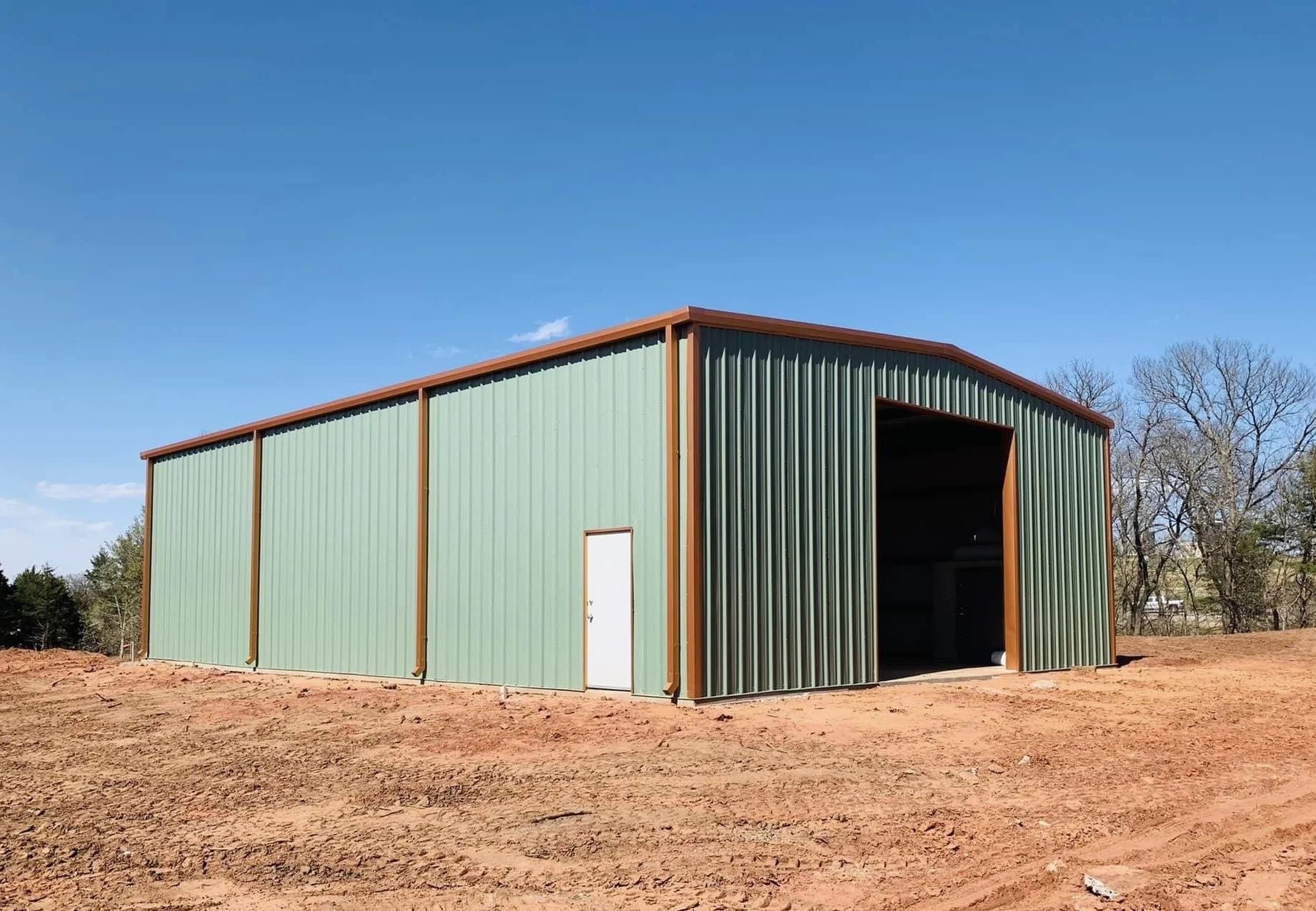 A large green metal storage building with a white door and an open bay, situated on a patch of dirt with trees in the background under a clear blue sky.