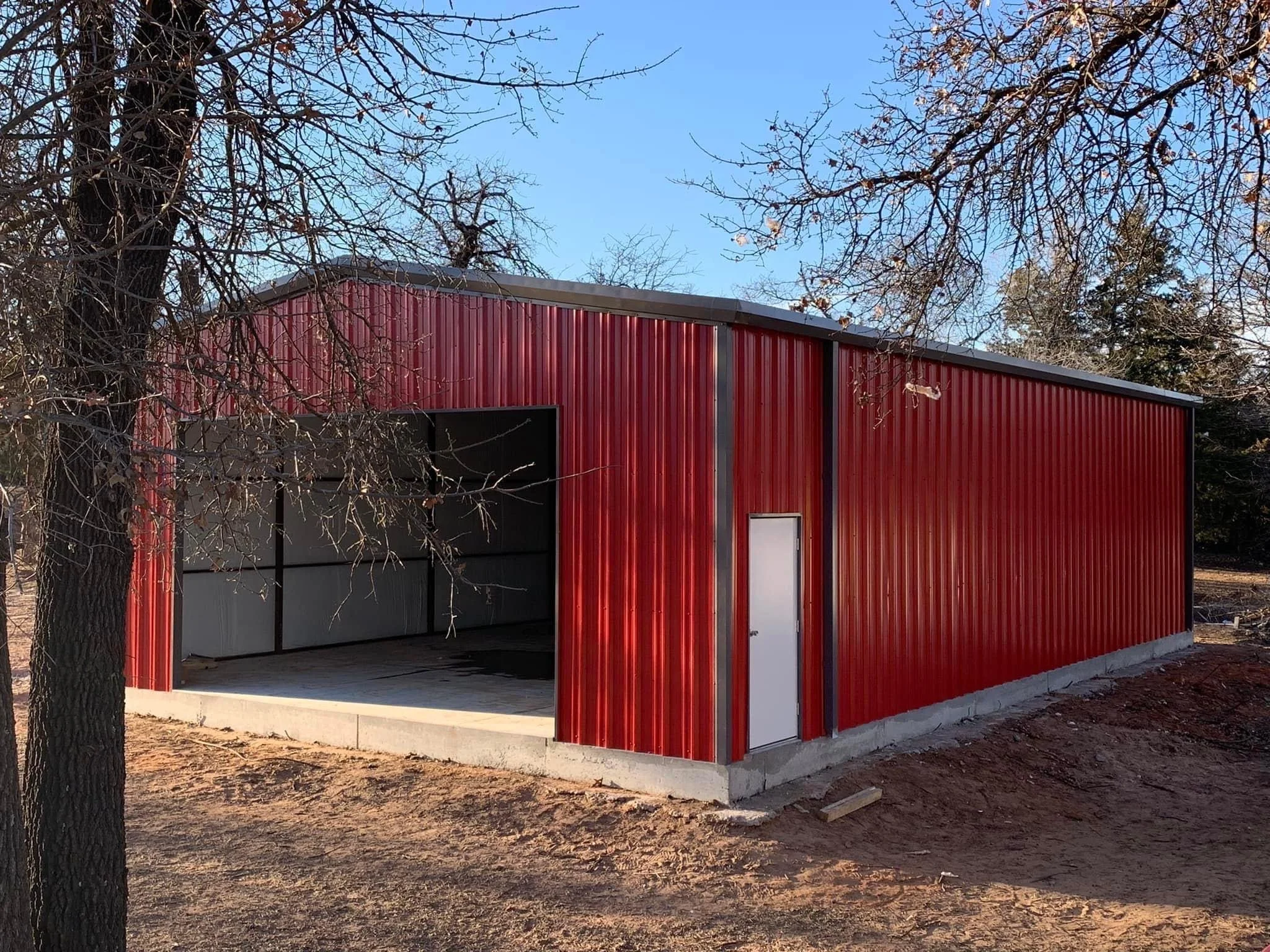 A red metal building with a small open garage door, surrounded by trees with bare branches, under a clear blue sky.