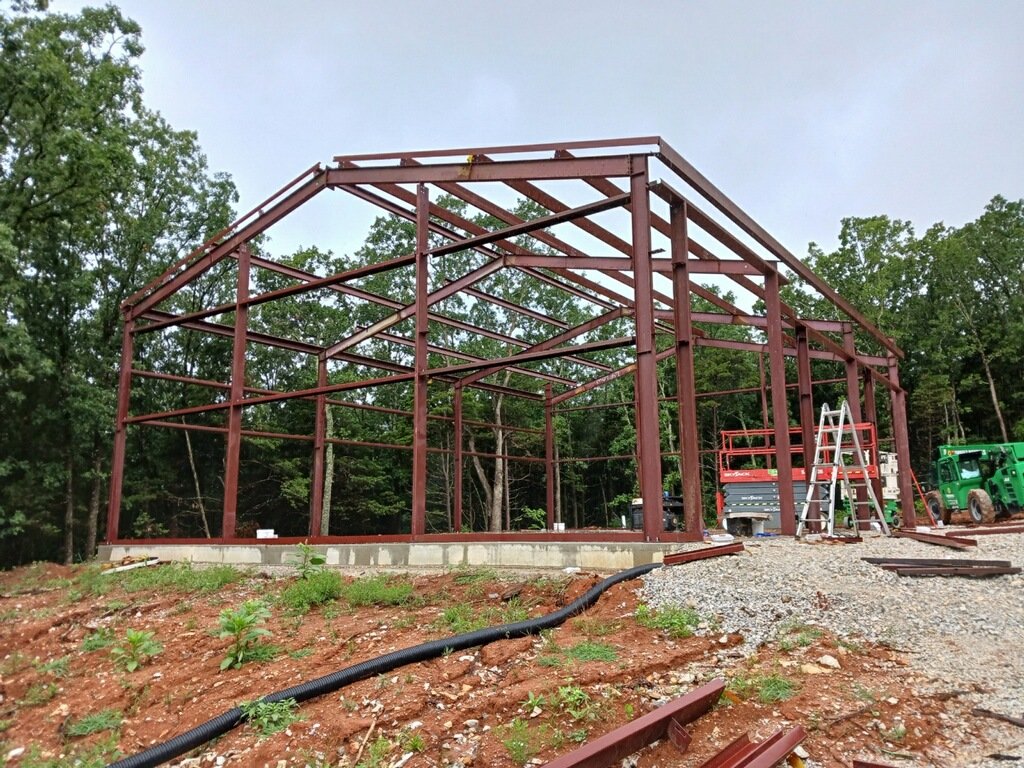 Steel frame construction of a building on a concrete foundation with construction equipment and ladders on site, surrounded by trees.