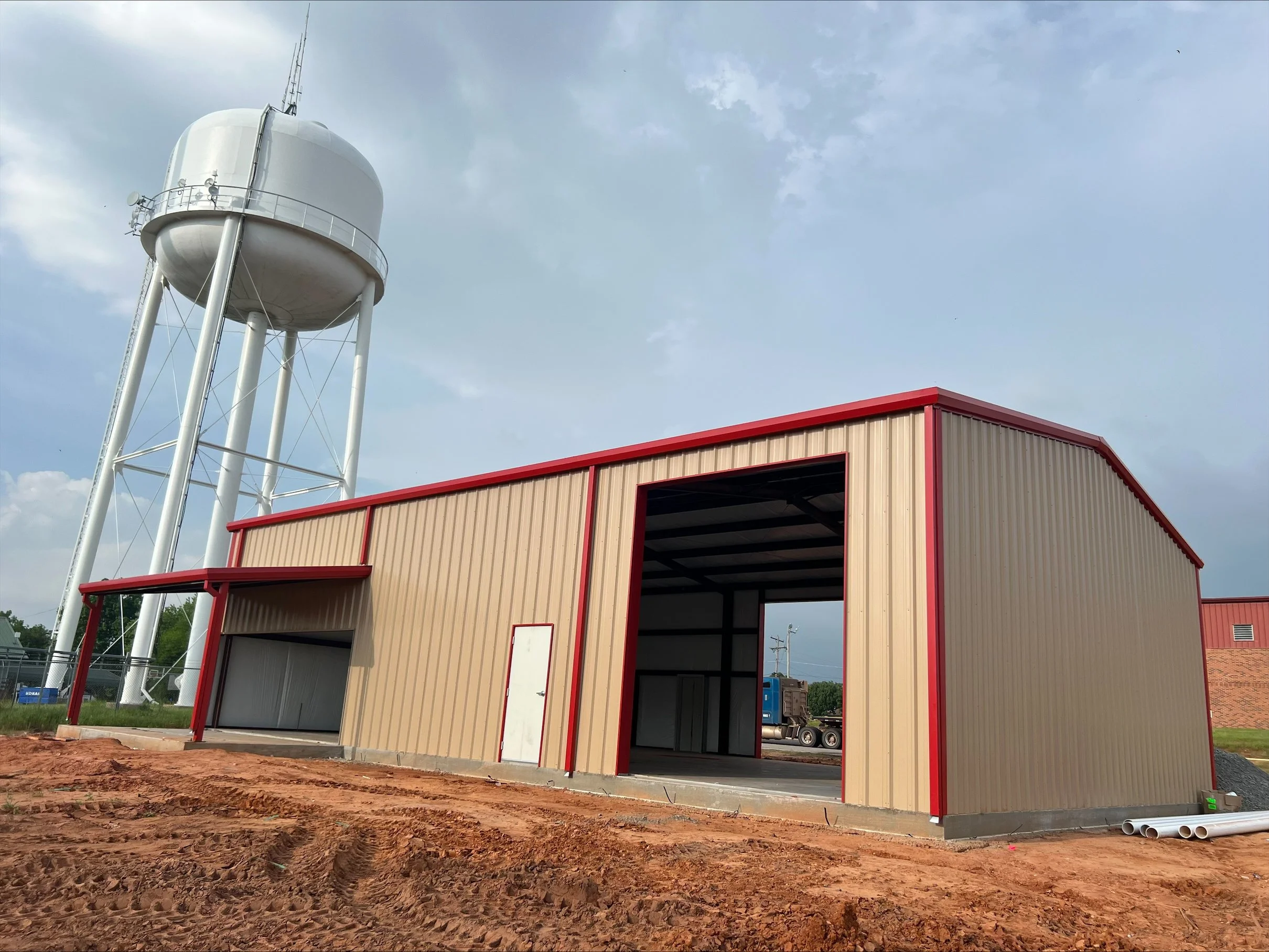 Construction site with a beige and red building structure and a large white water tower in the background under a cloudy sky.