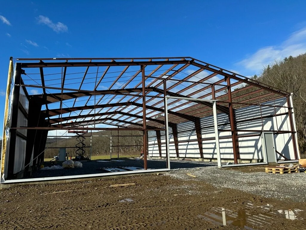 Construction of a metal-framed building or barn on a dirt lot with a blue sky and some trees in the background.