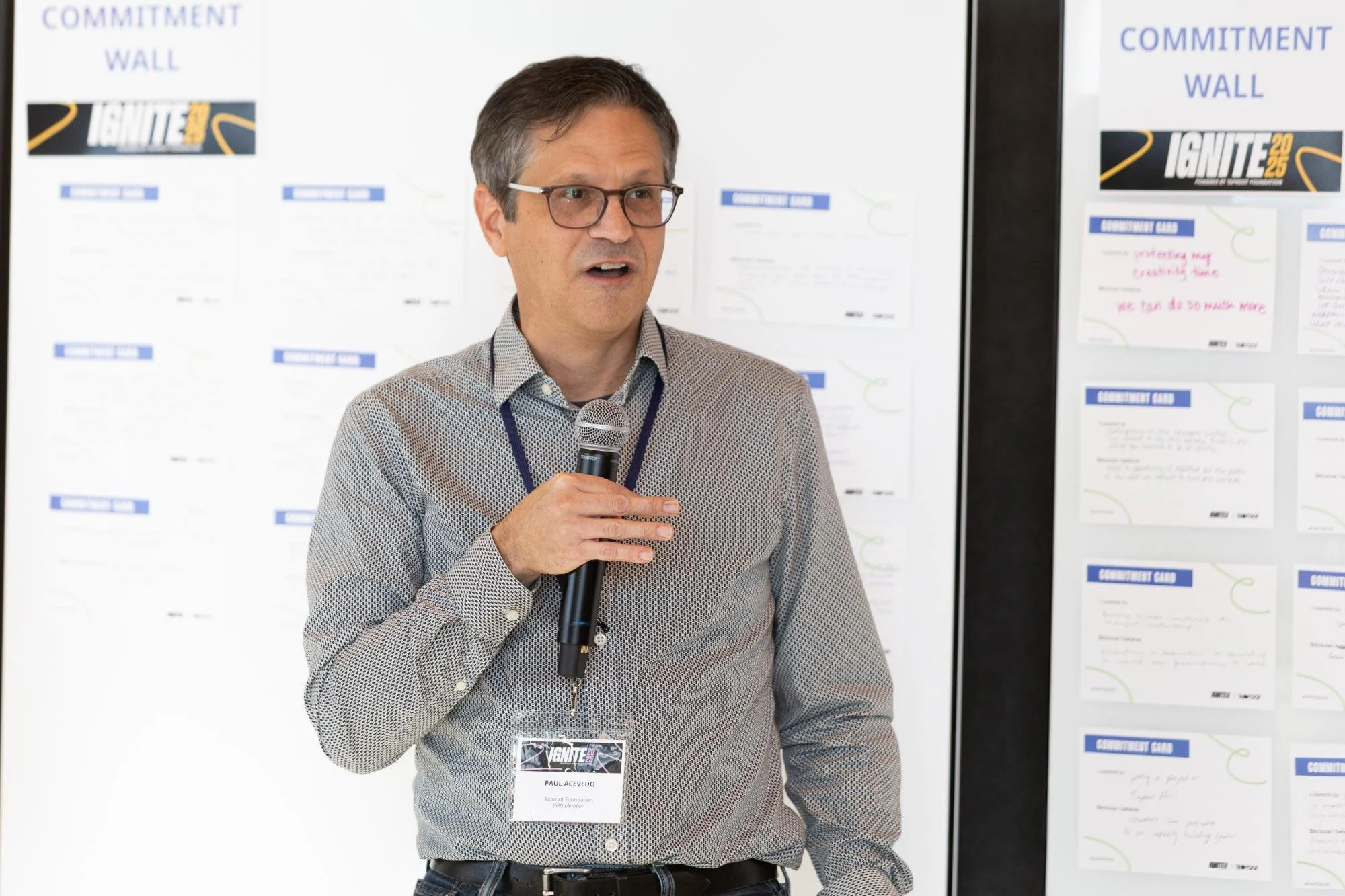 Man with glasses and a patterned dress shirt speaking into a microphone during an event, standing in front of a white display board with printed materials.