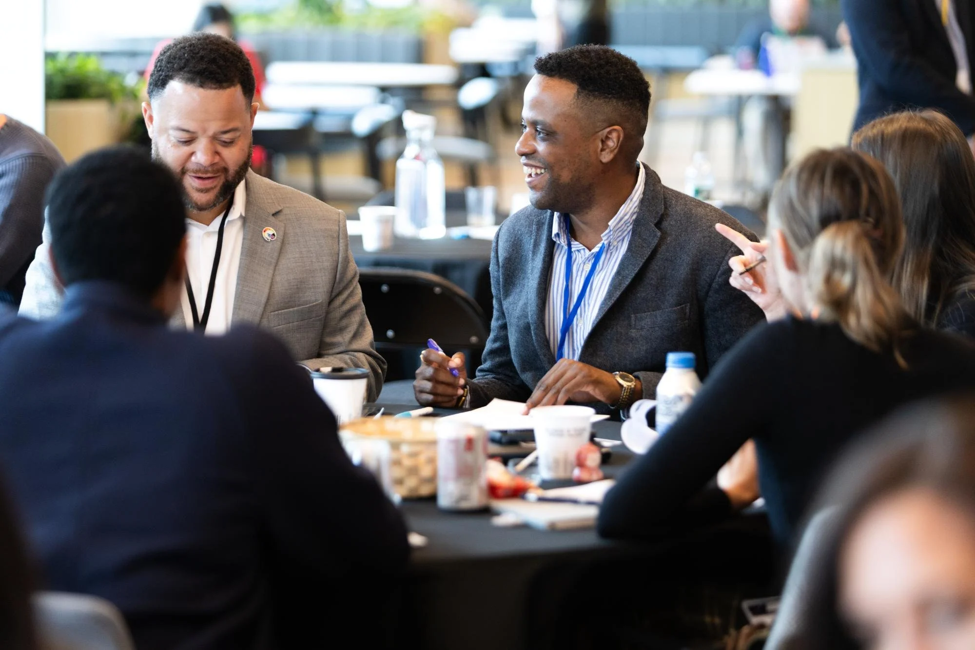 A group of people engaged in a discussion at a round table during a conference or workshop, with coffee cups and water bottles visible.