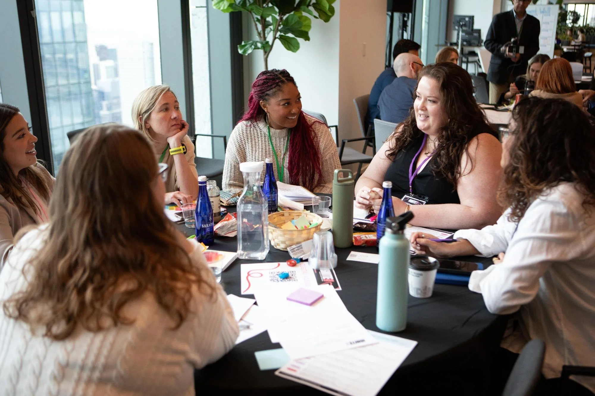 A group of women sitting around a conference table during a meeting or workshop, engaged in conversation, with water bottles, papers, and snacks on the table.