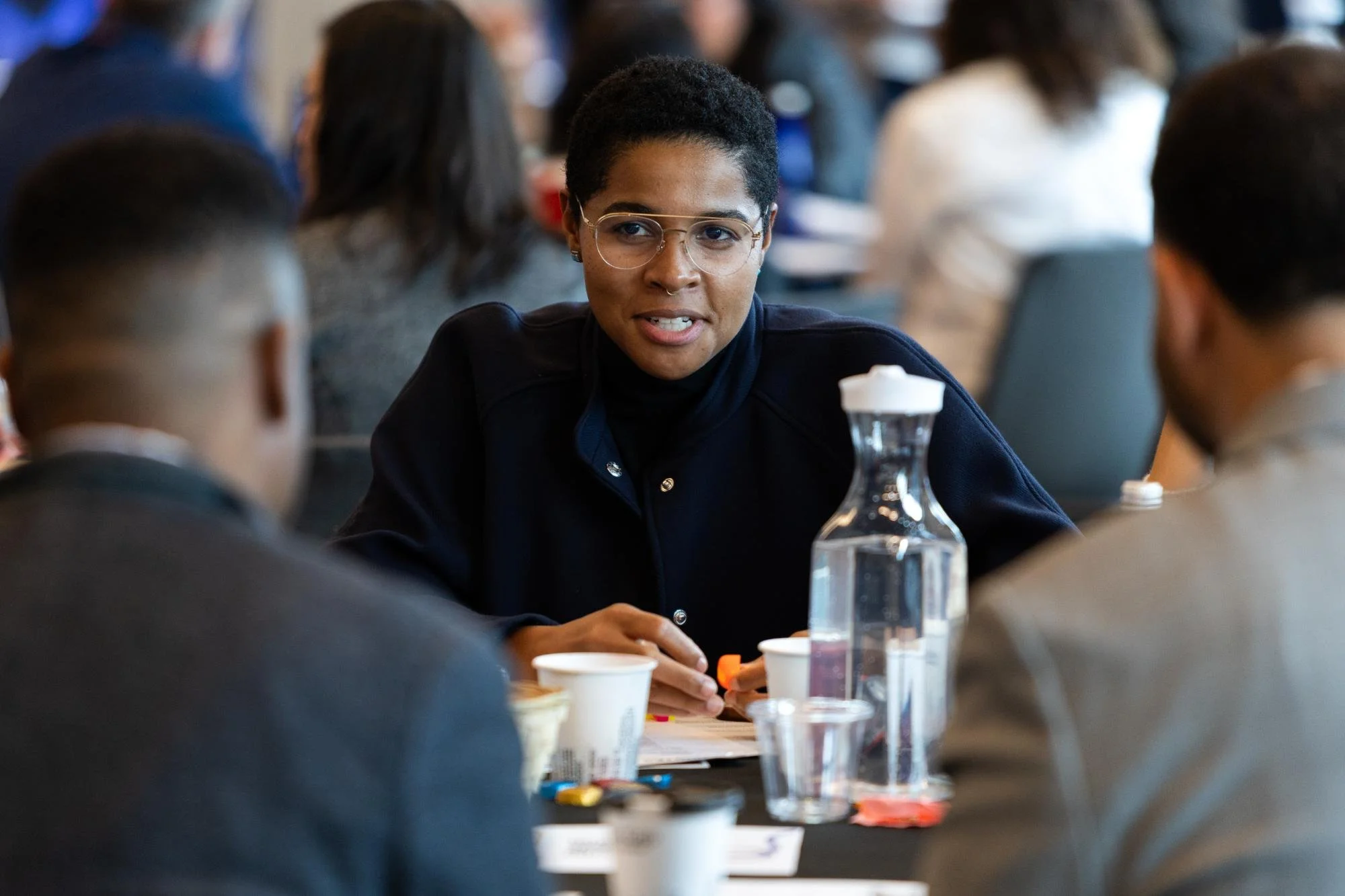 A woman with short curly black hair, glasses, and a dark shirt engaged in a discussion during a meeting, surrounded by other participants at a conference table with cups, water bottles, and papers.