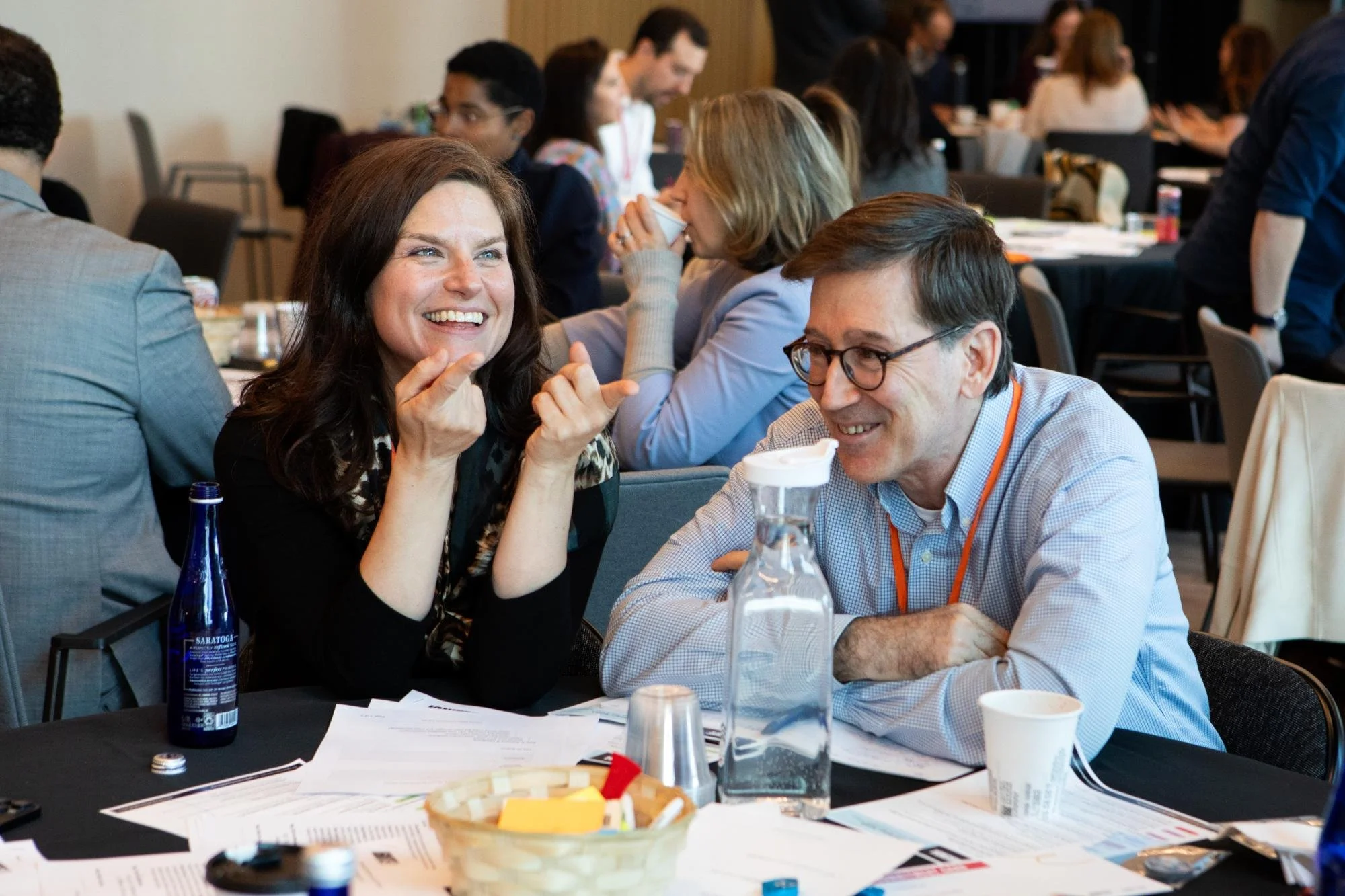 People sitting at tables in a conference room, engaging in conversation and smiling. Papers, pens, water bottles, and coffee cups are on the tables.