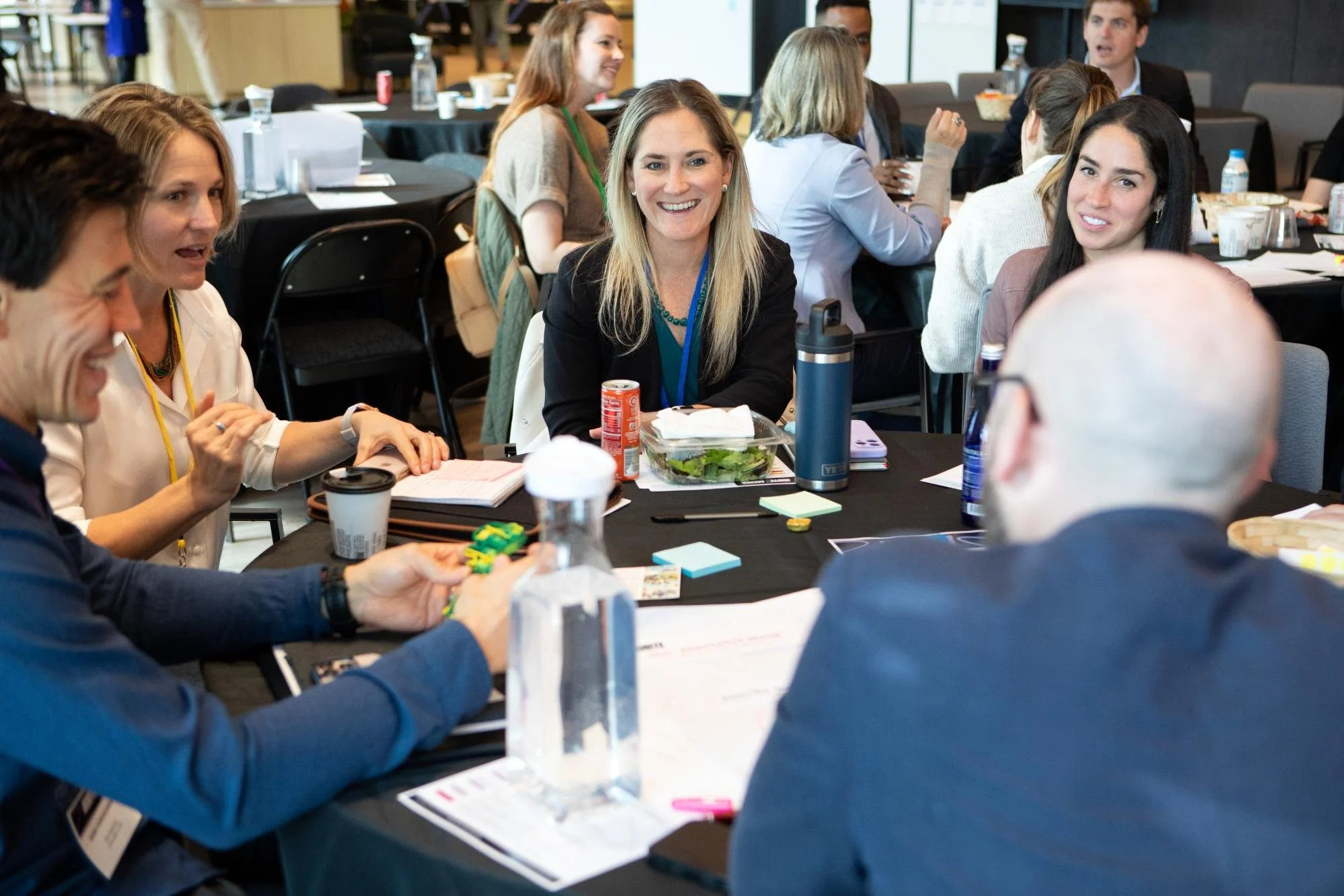 A group of diverse adults sitting at round tables in a conference room during a meeting, engaging in discussions and smiling.
