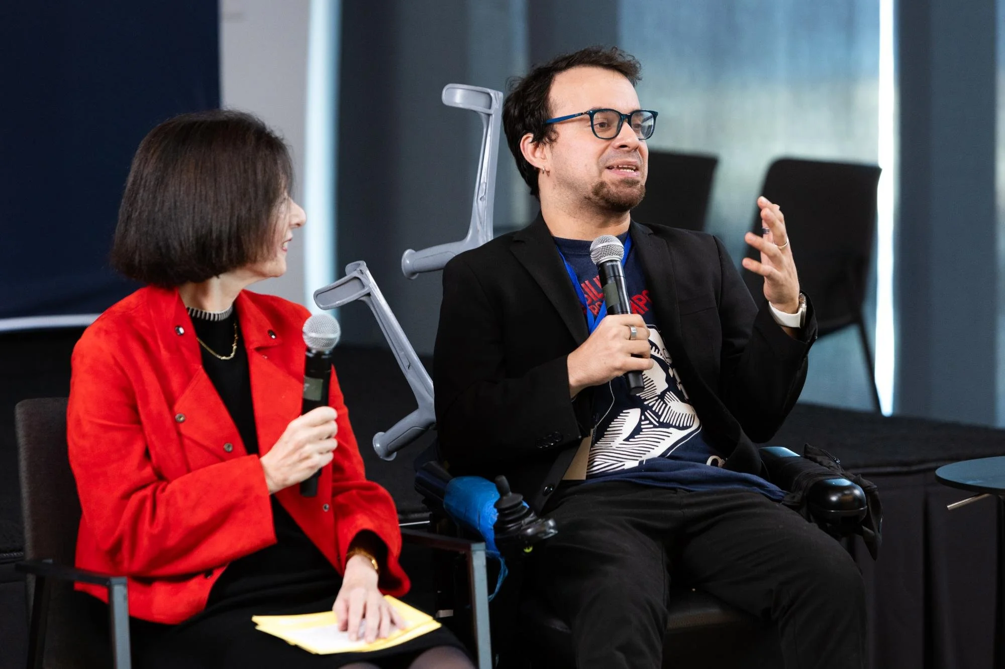 Two people seated, participating in a discussion. The person on the left is a woman with short dark hair, wearing a red blazer, holding a microphone and some yellow papers. The person on the right is a man in a wheelchair with dark hair and glasses, 