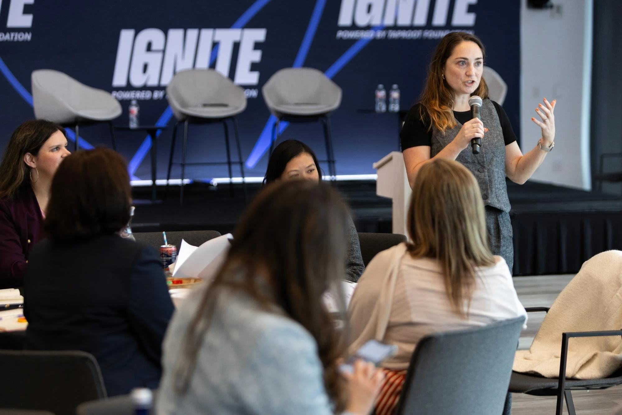 A woman with long brown hair speaking into a microphone during a panel presentation at a conference, with an audience of women seated around tables.
