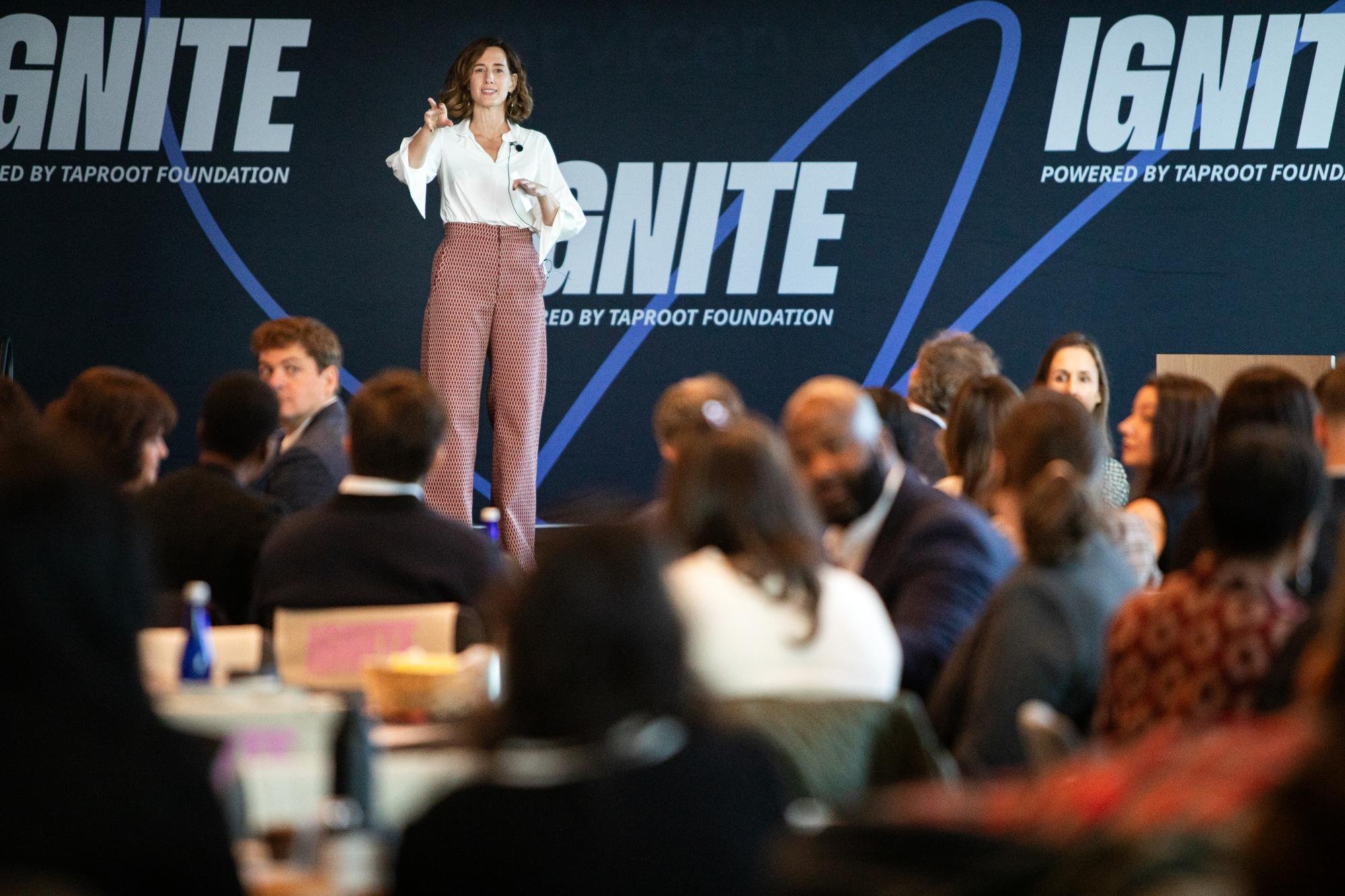 A woman speaking at a conference on a stage with a large background display featuring the words "SIGHT" and "IGNITE," with people seated in the audience.