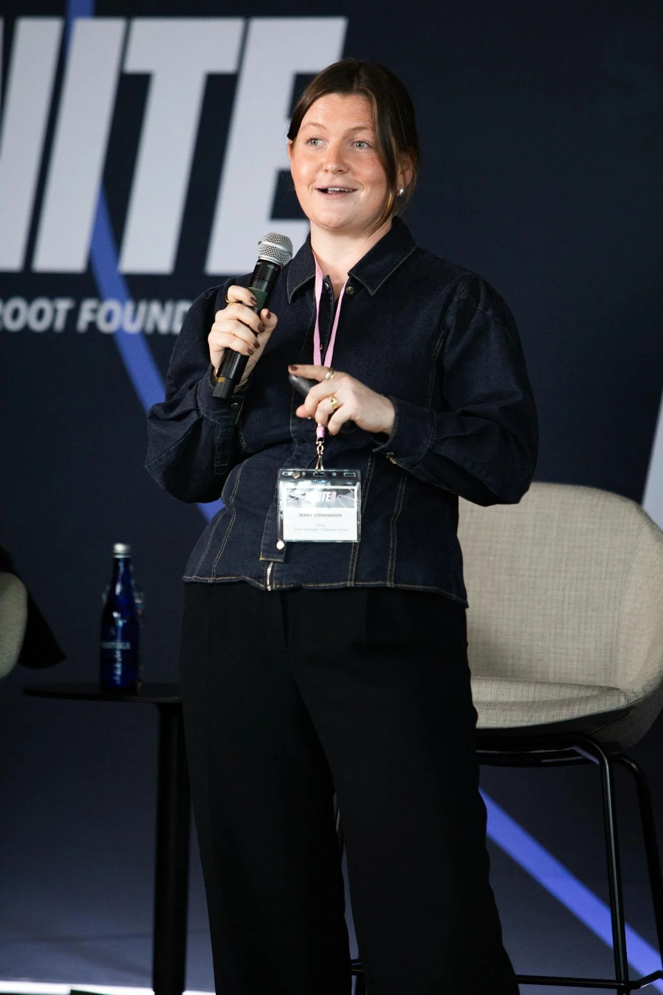 A woman with short brown hair and a black denim jacket holding a microphone, speaking at a conference or event with a black backdrop and a visible name tag around her neck.