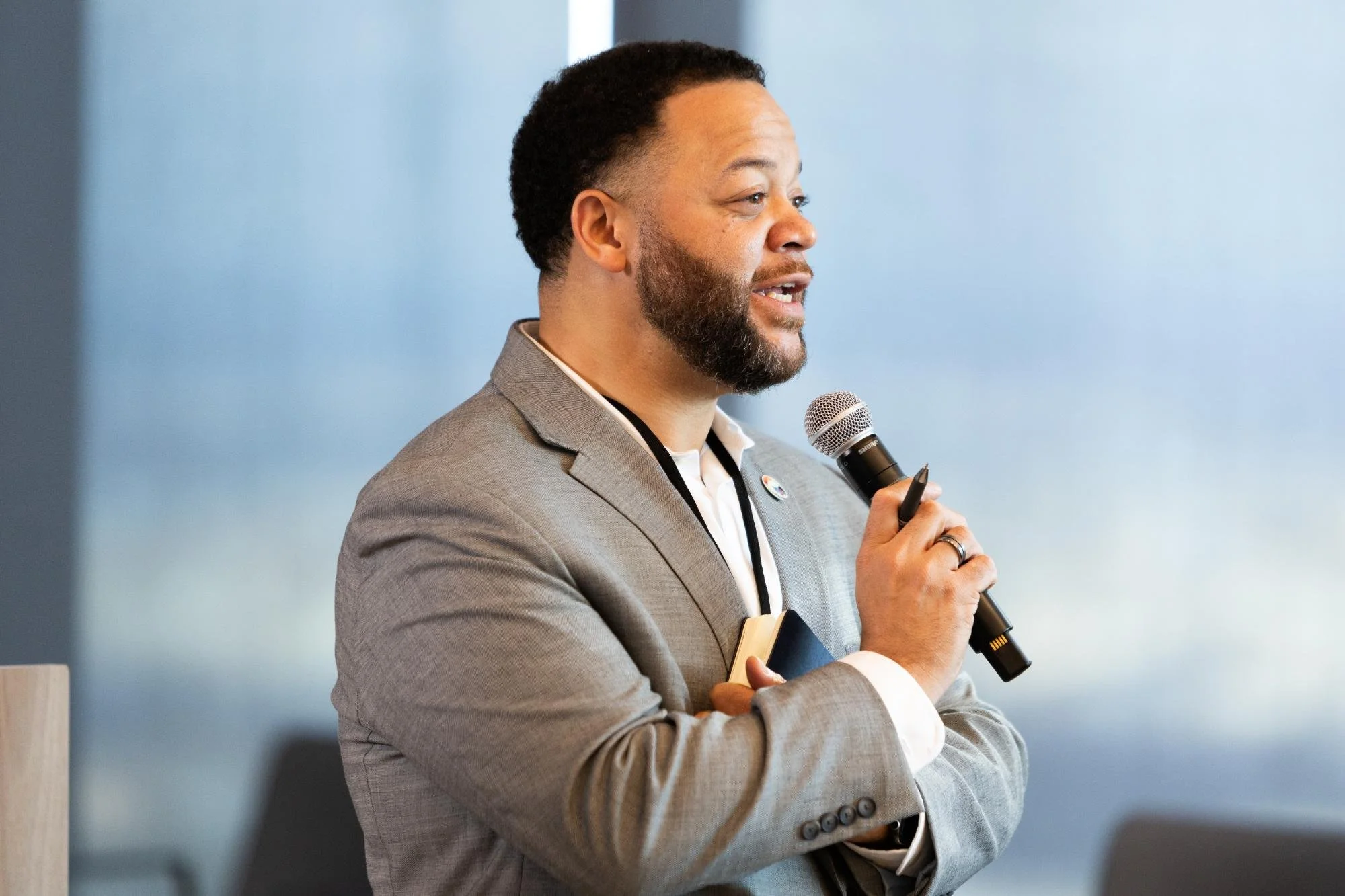 A man with a beard and mustache wearing a grey suit jacket and holding a microphone, speaking at an indoor event or conference against a blue wall background.