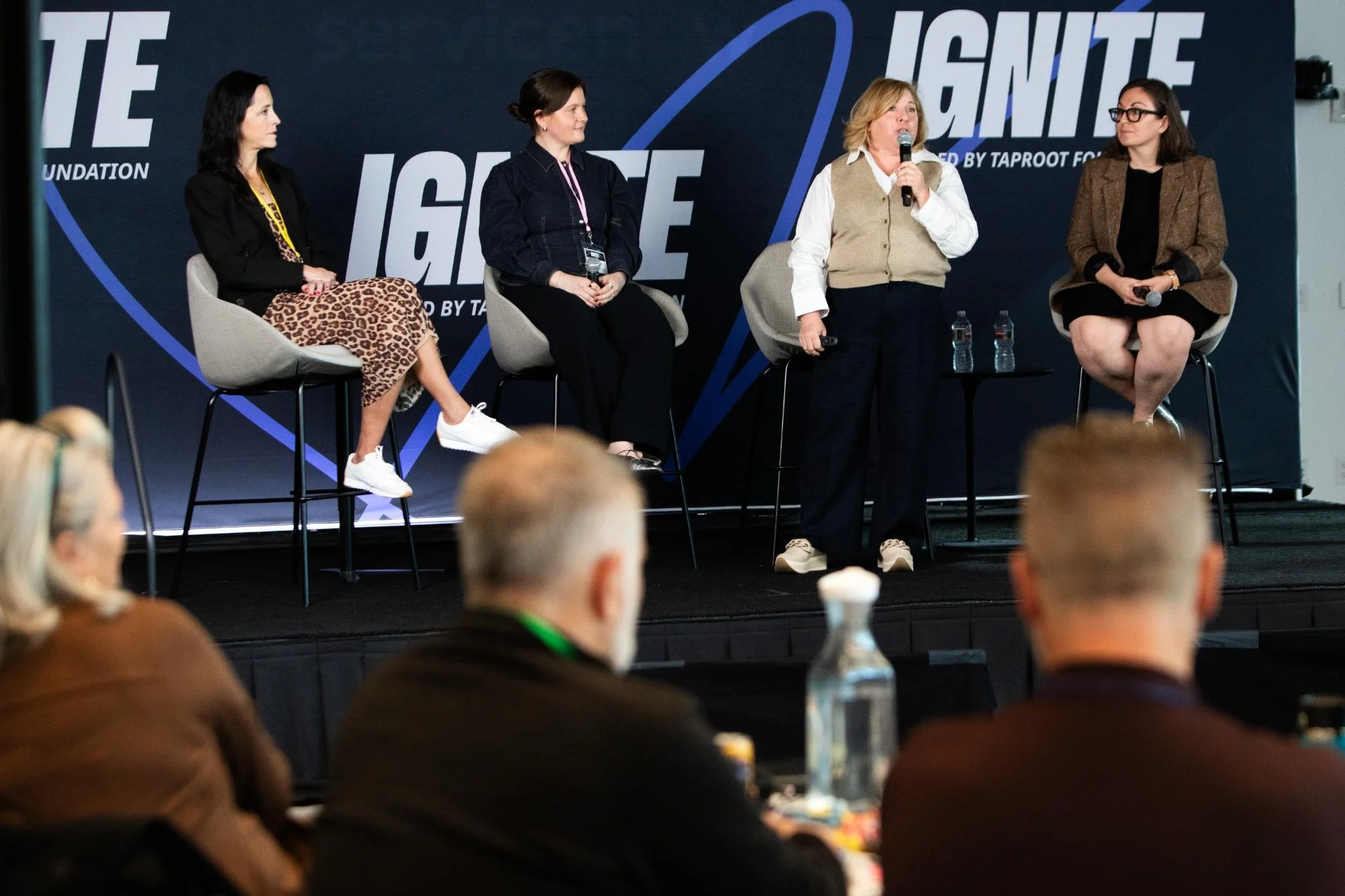 Four women seated on chairs on a stage, one standing and speaking into a microphone during a panel discussion at an event. Behind them, a large sign displays the words 'Ignite' and related text. Audience members are visible in the foreground, blurred