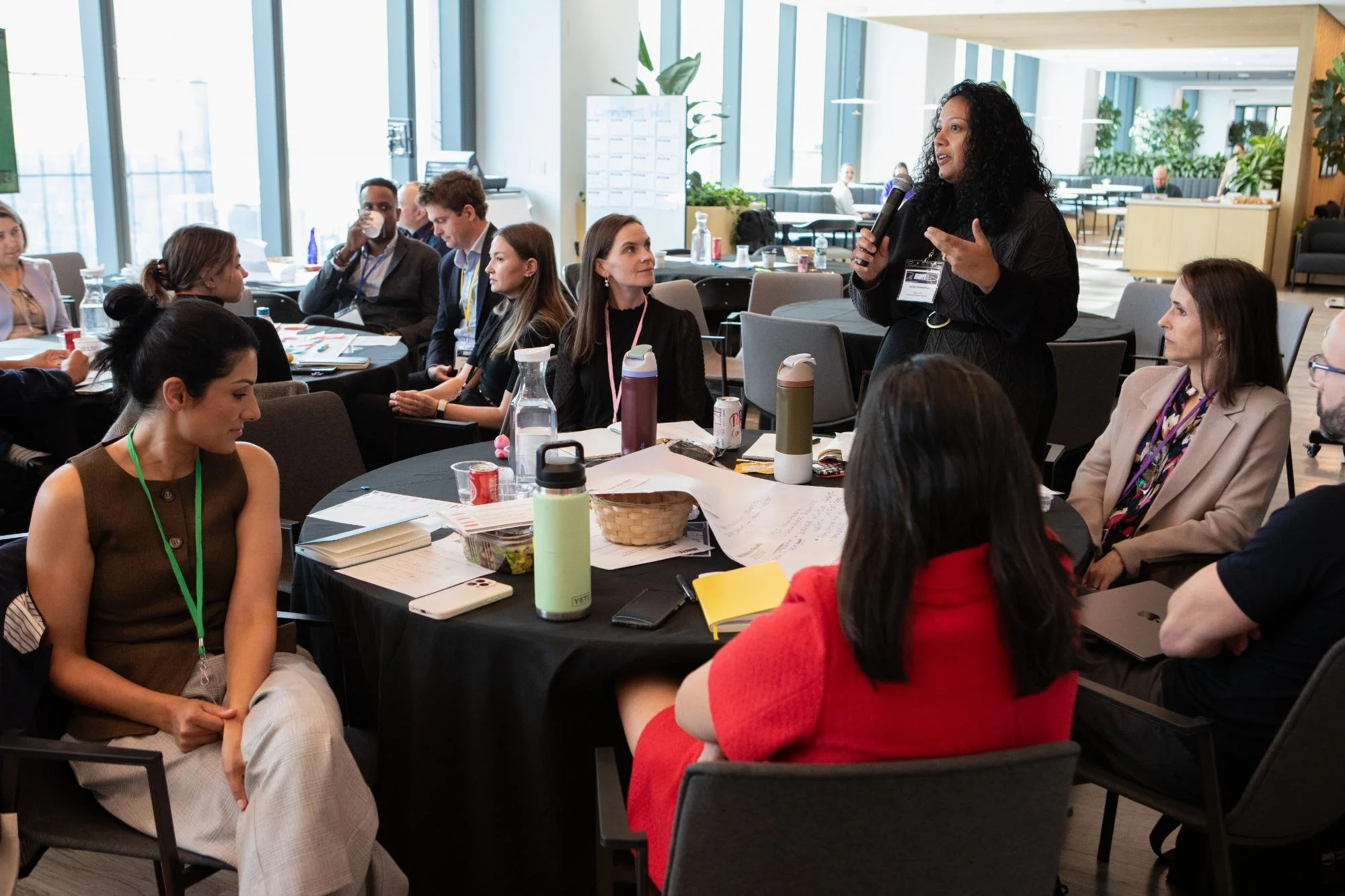 A woman speaking into a microphone during a meeting or conference in a modern, well-lit office space with large windows, surrounded by attendees seated at round tables.