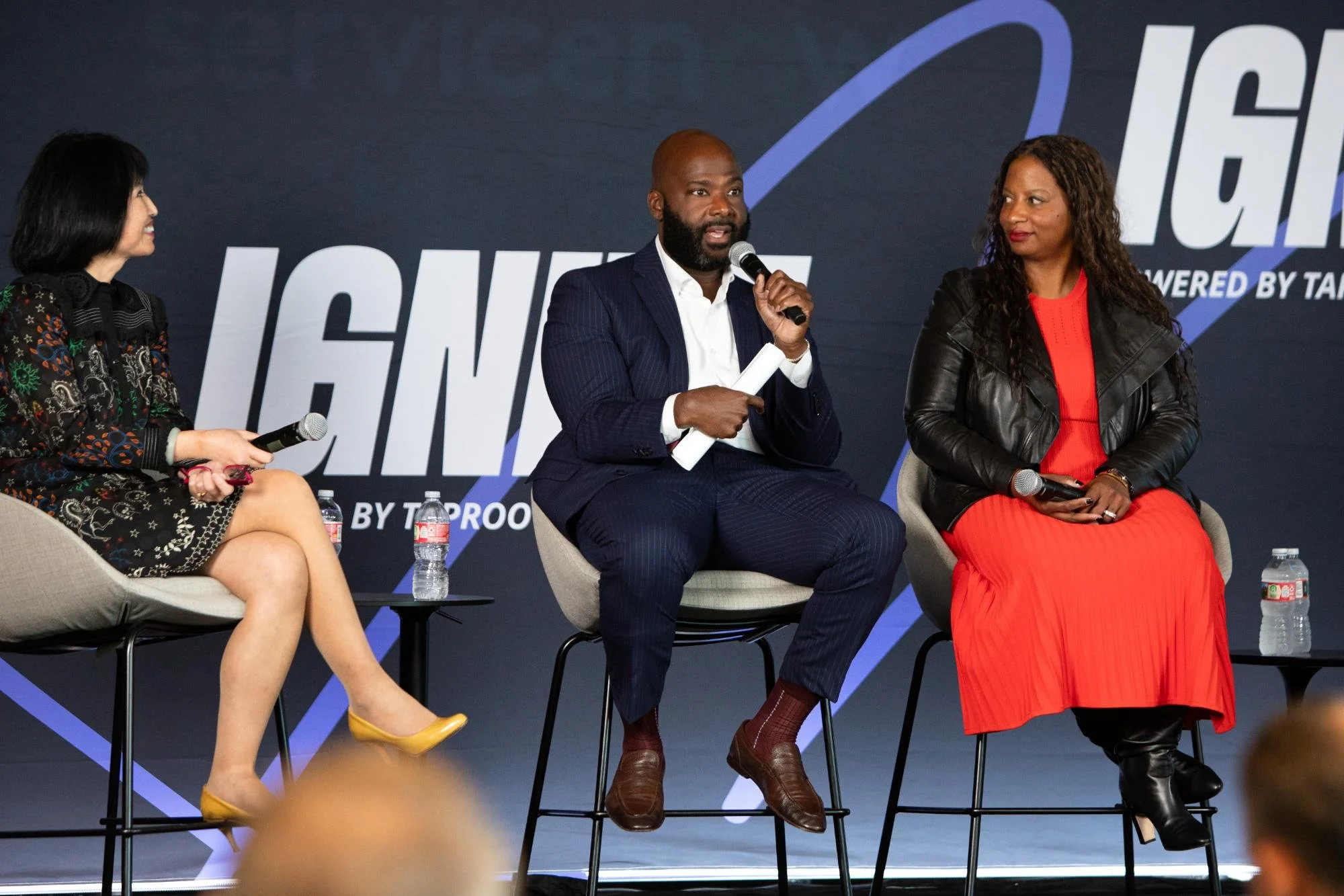 Three people sitting on stage during a panel discussion, with a blue backdrop and water bottles on small tables in front of them.