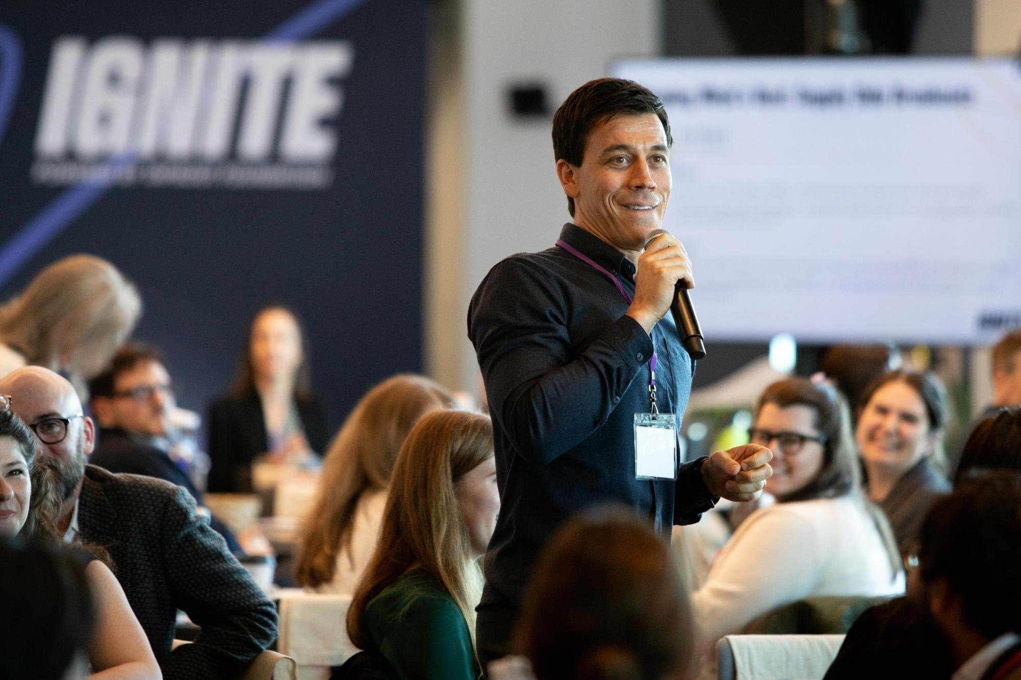 A man holding a microphone speaking at a conference with an audience seated around him. The background shows a large monitor and a banner with the word 'HEIGHTS' on it.