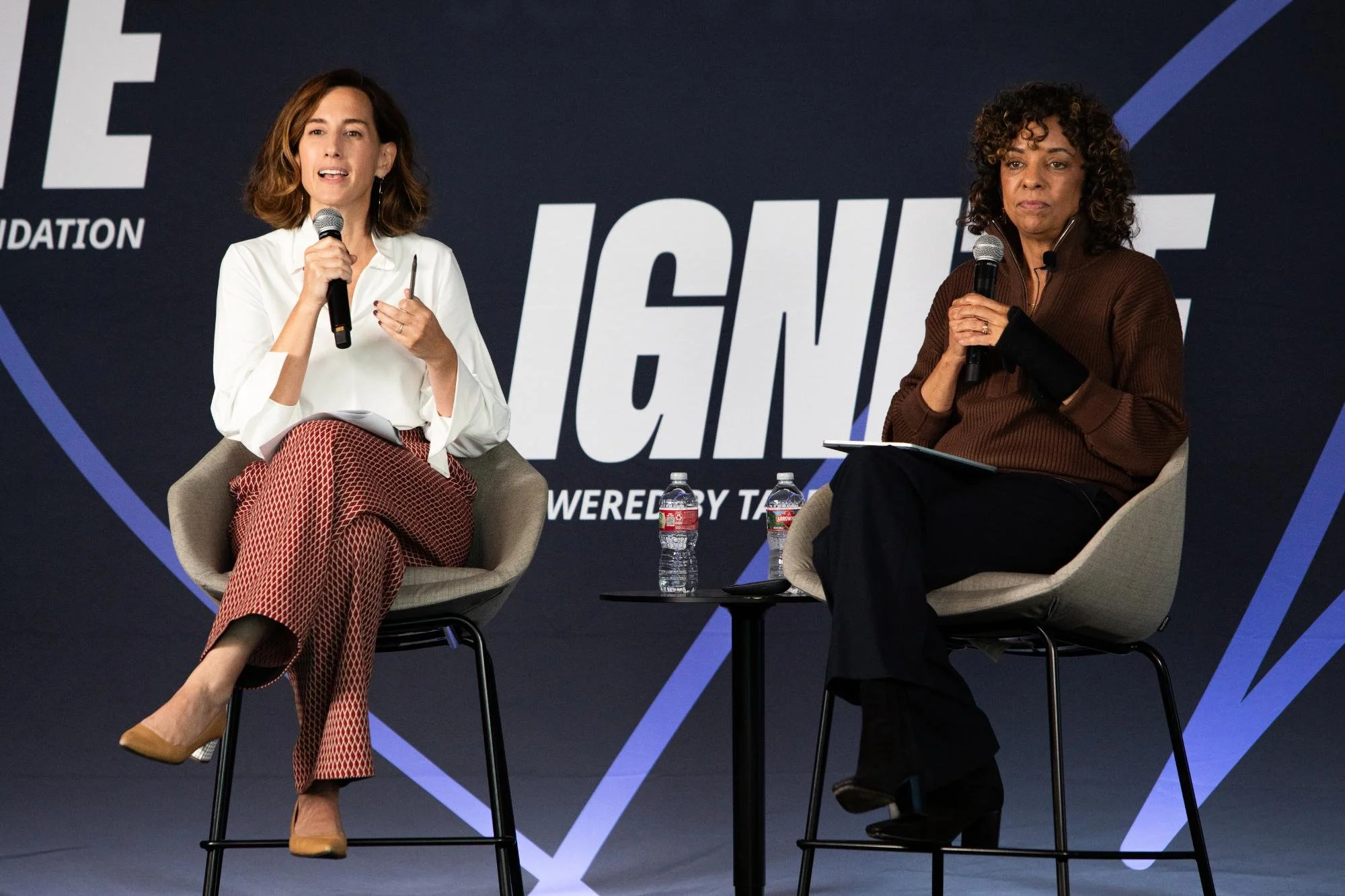 Two women seated on stage participating in a discussion, holding microphones with water bottles on a small table between them, behind them is a large dark background with partially visible text.