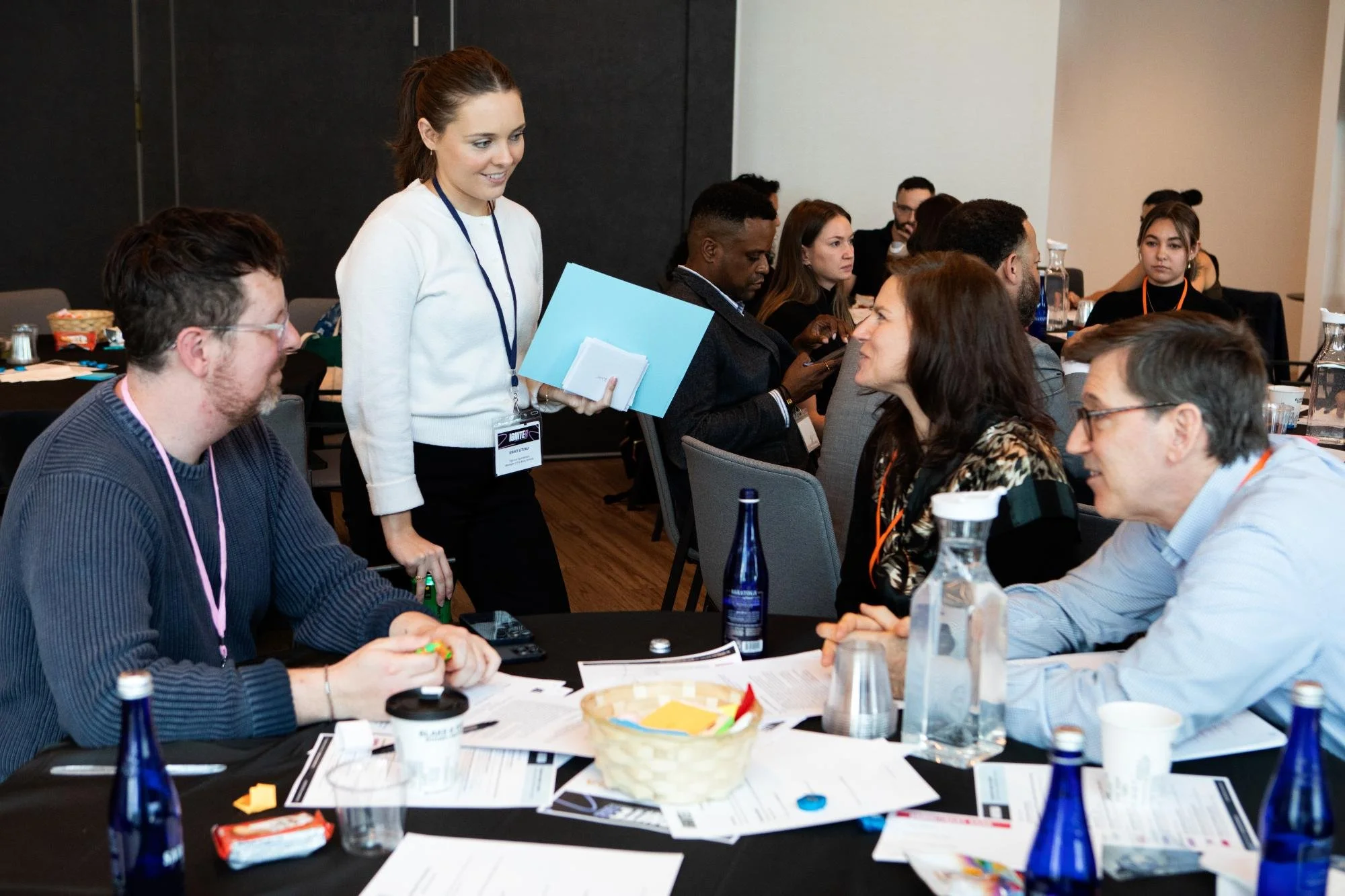 A group of diverse people attending a conference or workshop, seated at tables with documents, water bottles, and snacks, while a woman stands and engages with others.