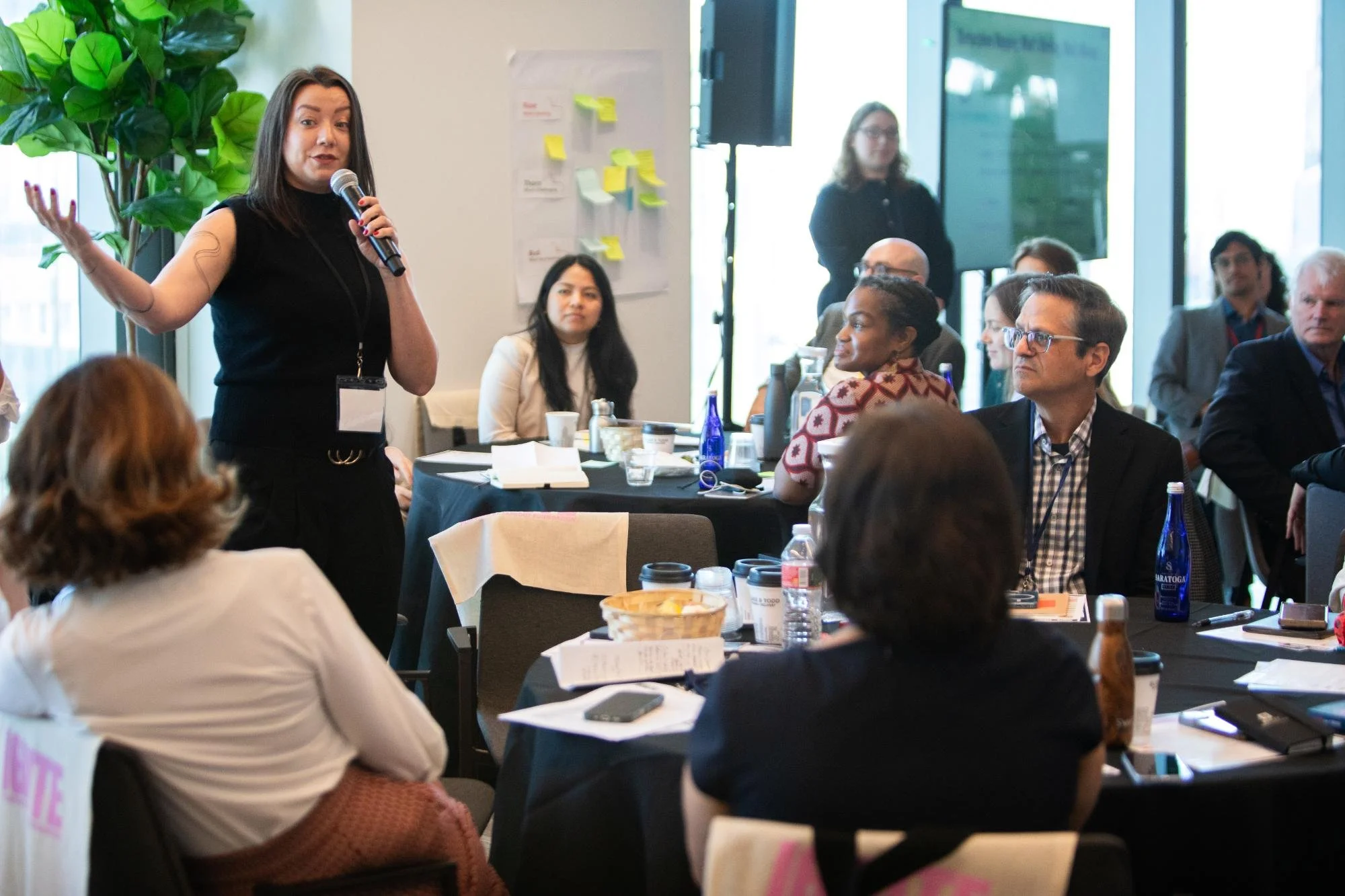 A woman with brown hair, wearing a black sleeveless top, is speaking into a microphone while gesturing with her left hand at a conference or meeting. Multiple attendees are seated around tables with notebooks, water bottles, and cups. Some are paying