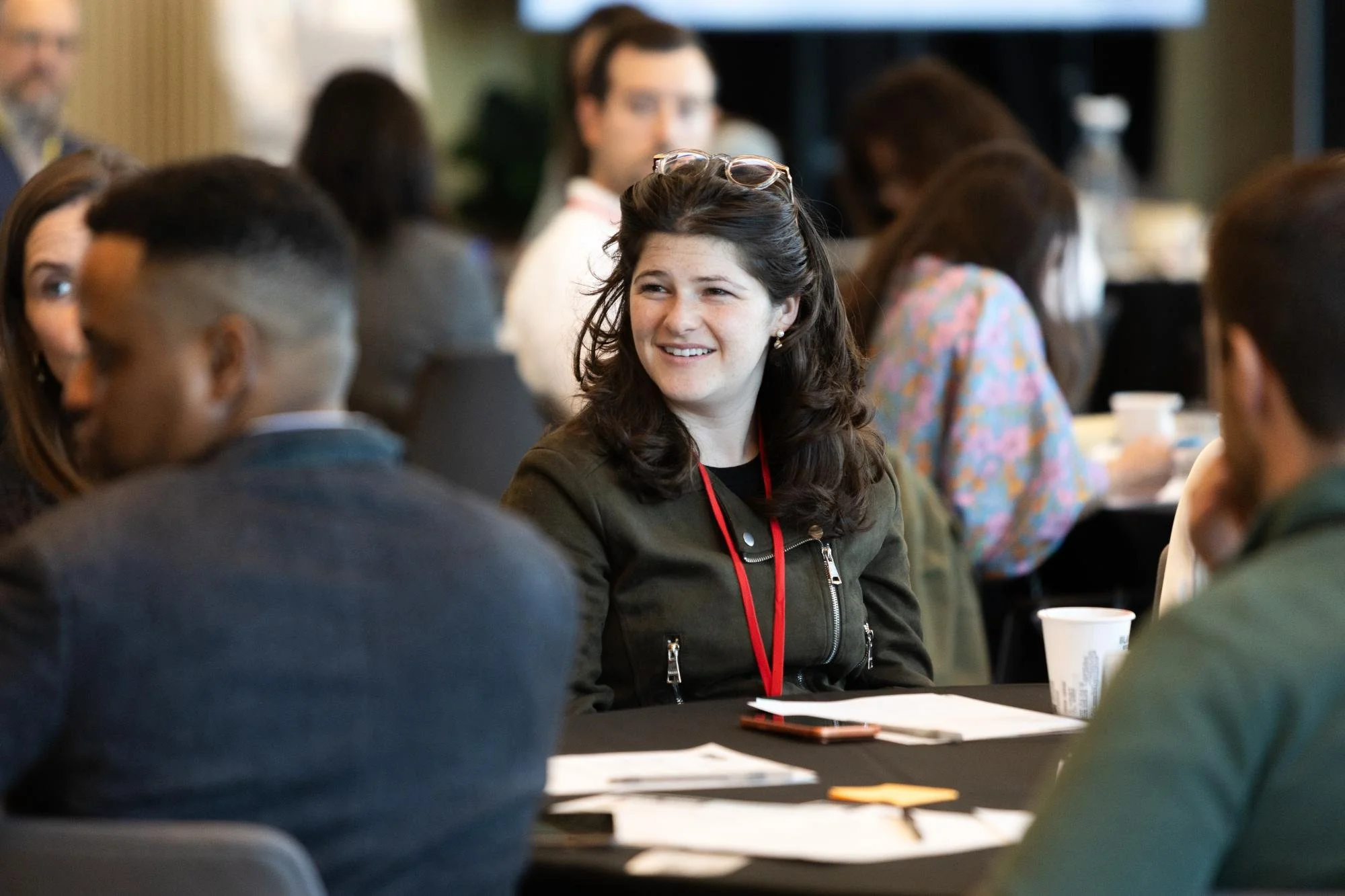 A group of people sitting around tables at a conference or meeting, with a woman smiling and wearing glasses on her head in focus.