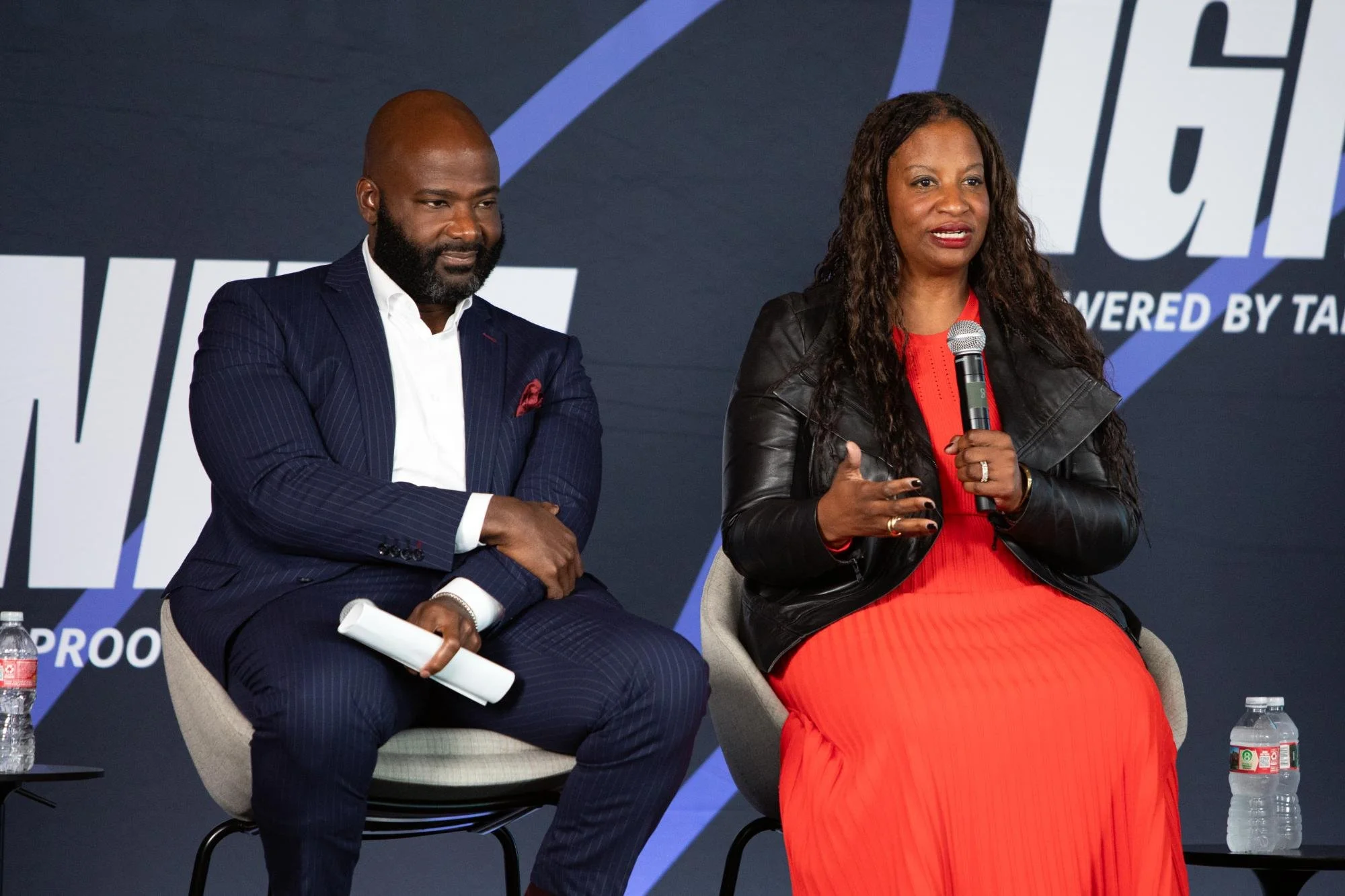 A man in a navy suit and a woman in a red dress and black leather jacket sitting on stage during a panel discussion. The woman is speaking into a microphone while the man looks on, with a dark background and a large partial logo or text behind them. 