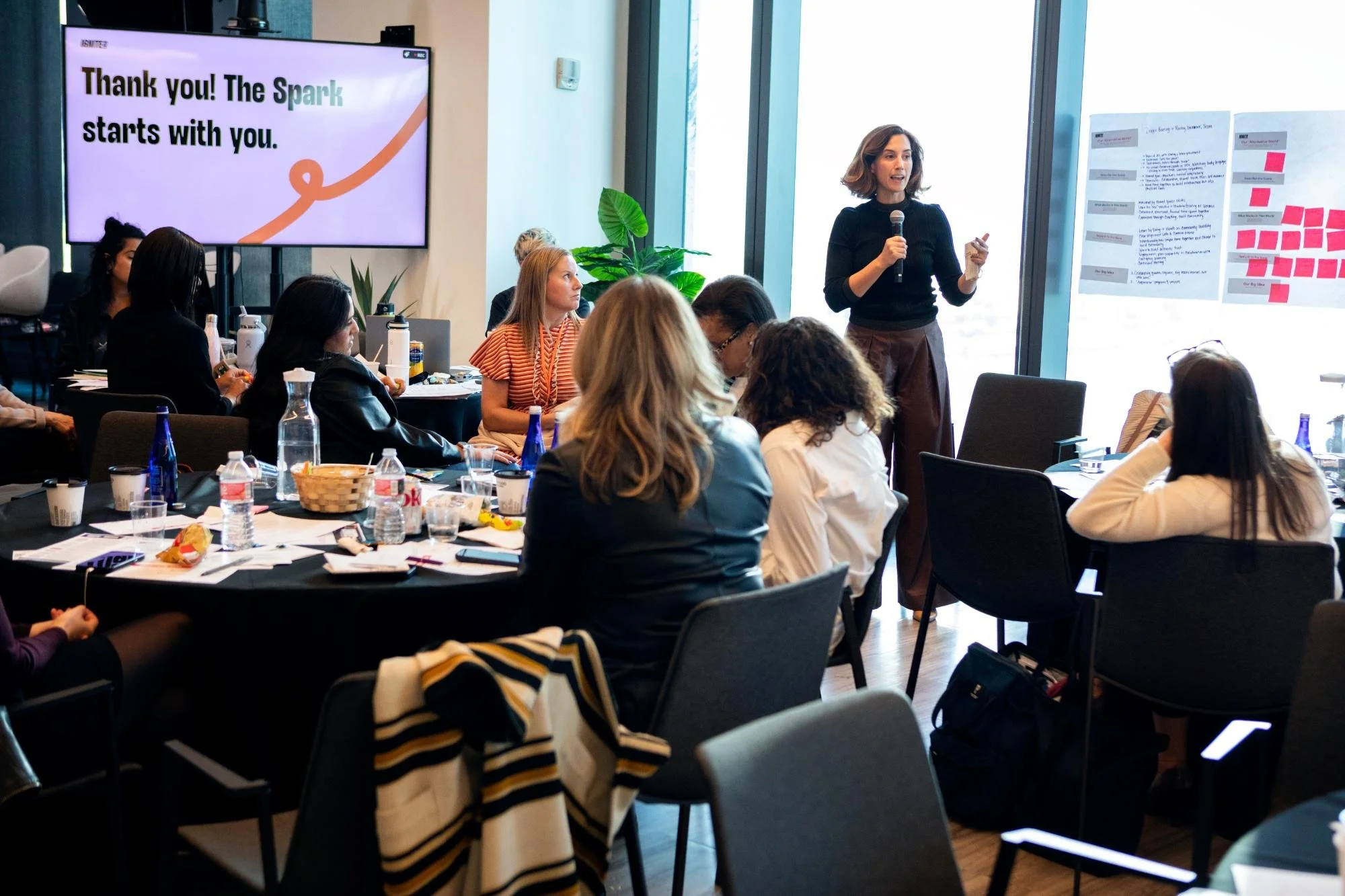 A woman standing and giving a presentation to an audience in a conference room with large windows. The presentation slide on the screen reads 'Thank you! The Spark starts with you.' Several people are seated at tables, listening and taking notes.