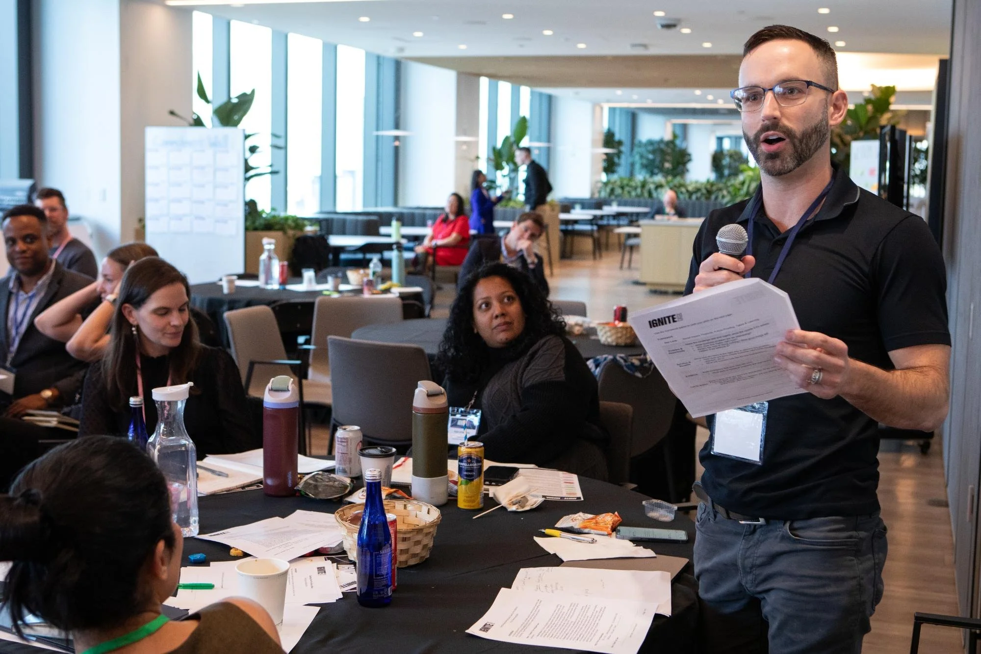 A man with glasses and a beard speaks into a microphone while holding a paper at a conference. Several people sit at a table, some looking at him, with drinks and papers on the table. The background shows a bright, modern conference space with large 