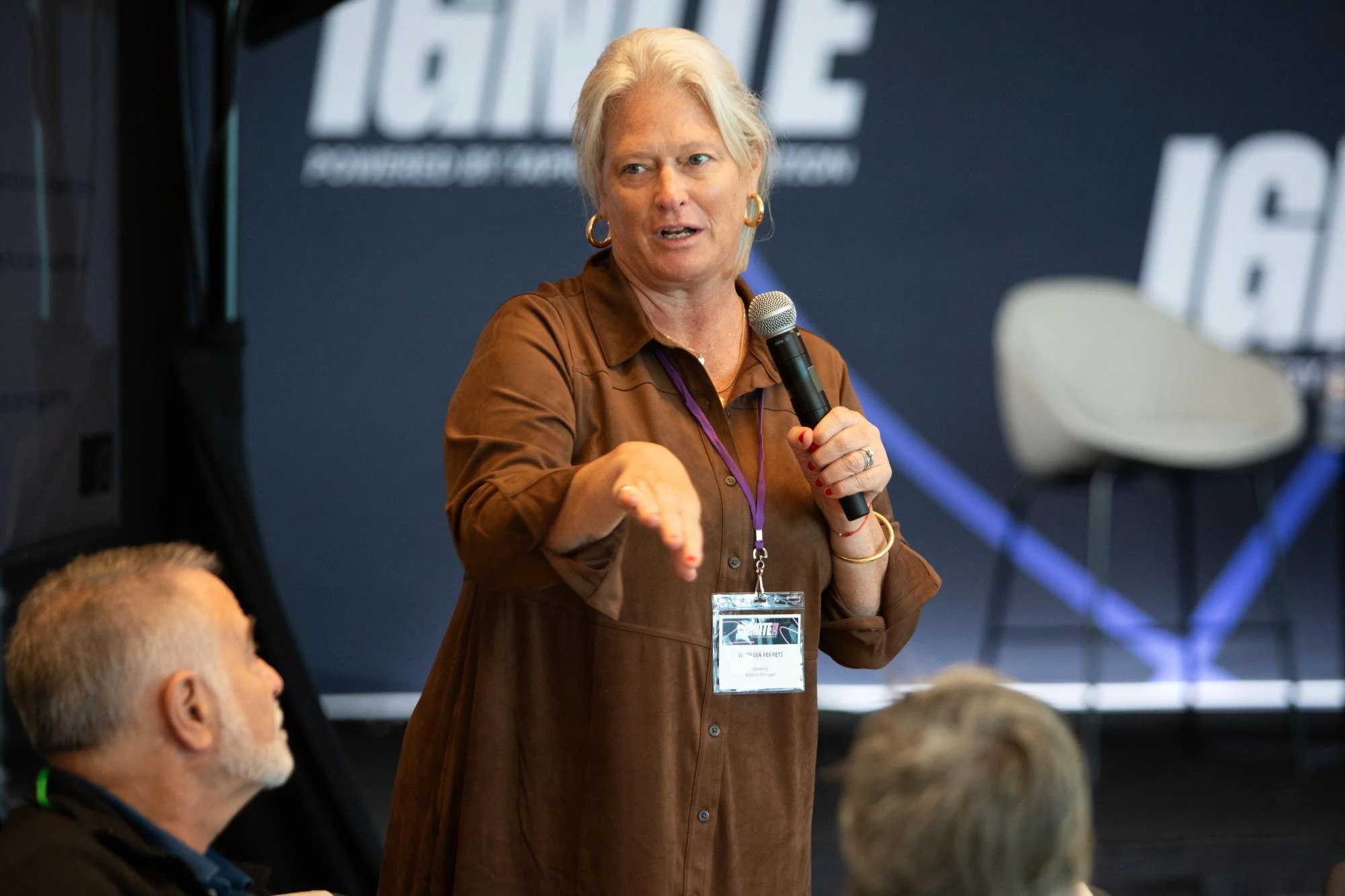A woman with gray hair, wearing a brown shirt and gold earrings, holding a microphone and speaking at a conference or meeting. Several people are seated around her, and there is a large screen or backdrop with text in the background.