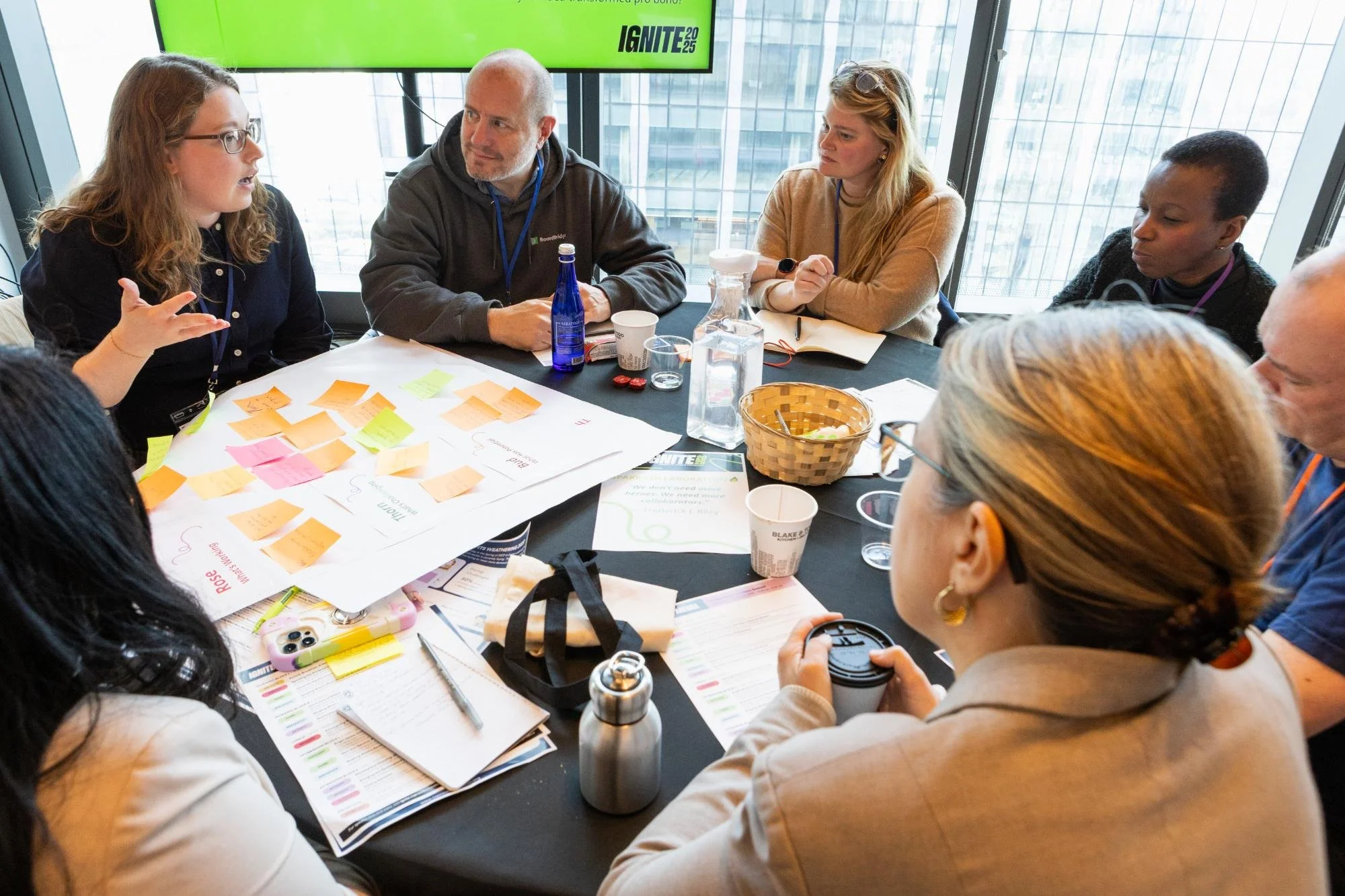 A group of diverse people sitting around a table engaged in a discussion, with sticky notes, papers, and notebooks on the table, in a bright conference room with large windows.