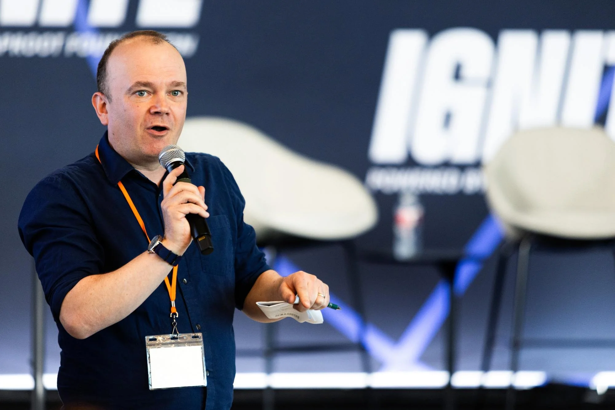 A man holding a microphone and speaking at a conference or event. He is wearing a dark blue shirt and has a notepad or paper in his other hand. In the background, there are blurred images of chairs and a large screen with the word 'LIVE' partially vi
