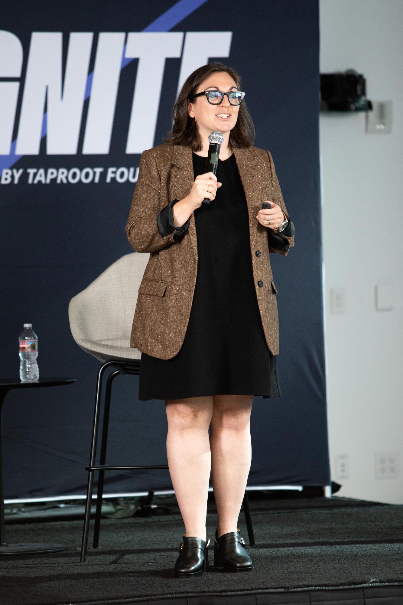 A woman with brown hair, glasses, and pale skin is speaking into a microphone while wearing a brown blazer, black dress, and black shoes. She is standing on a stage with a backdrop displaying the word 'VOTE' in large letters.
