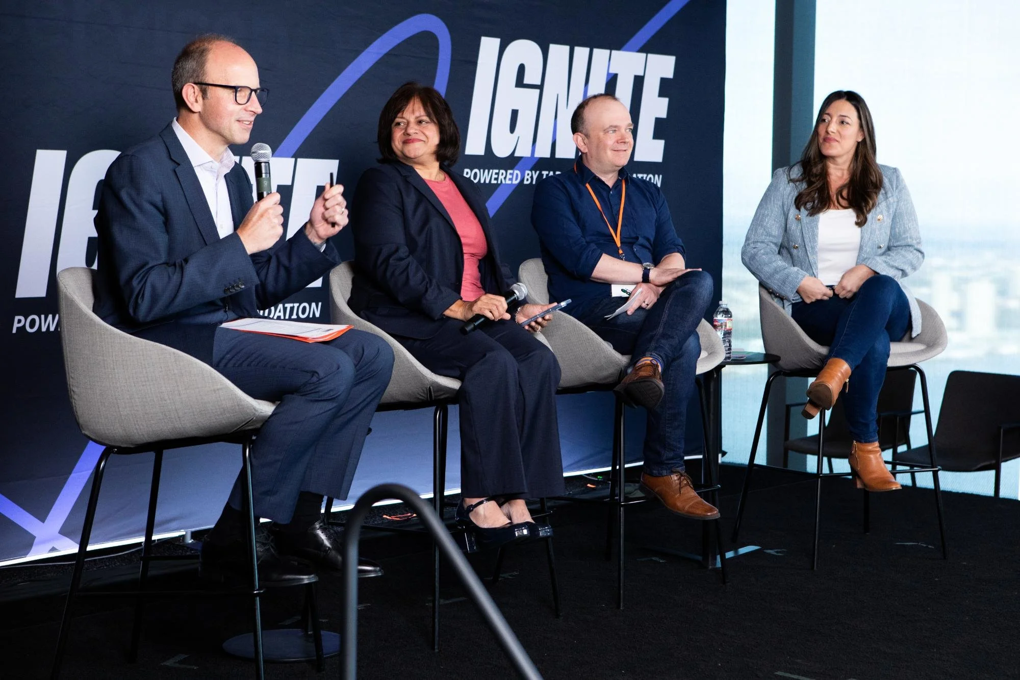 Four individuals sitting on stage at a panel discussion with a backdrop reading "Ignite." The first person, a man, is speaking into a microphone and wearing glasses, a suit, and a white shirt. The second person, a woman, has dark hair and is wearing 
