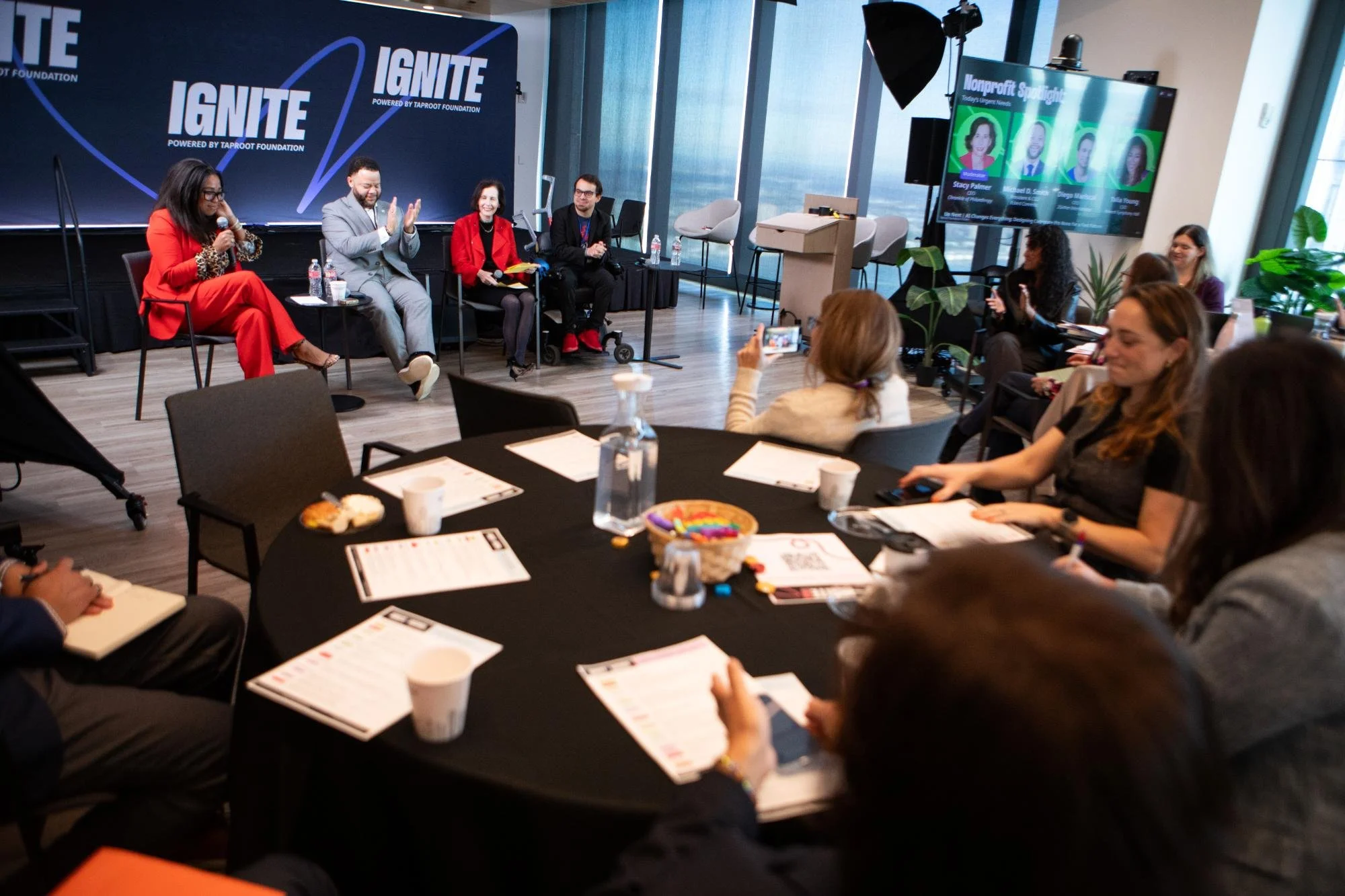 Panel discussion at an event with four speakers sitting on stage facing an audience. The event is called IGNITE, powered by the Taproot Foundation, as shown on the large backdrop behind the speakers. Attendees in the audience are seated at round tabl