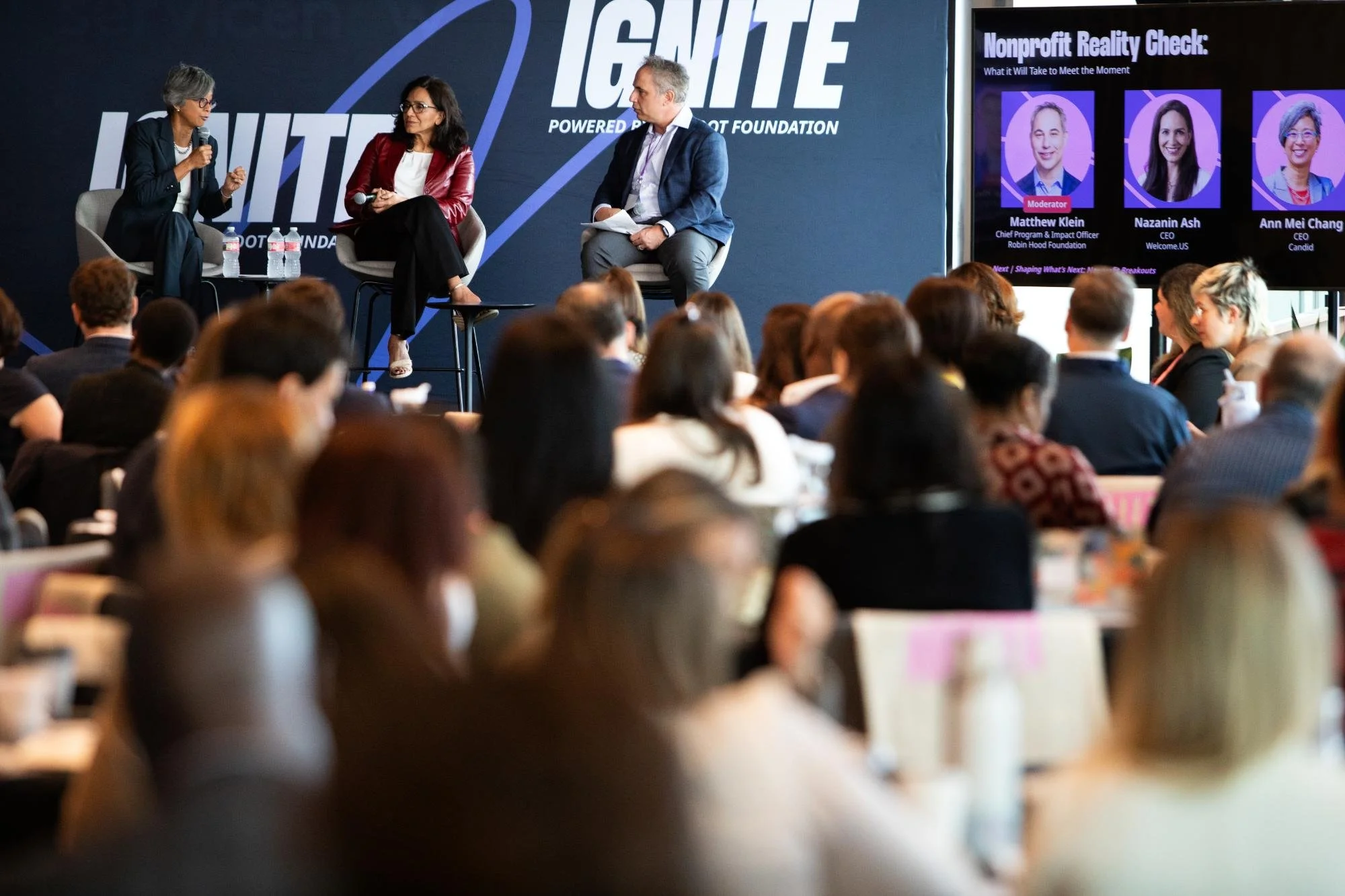 A panel discussion at a conference with three people on stage and a large audience. A woman on the left is speaking into a microphone, sitting with water bottles in front of her. The middle woman and man are listening. There is a big screen to the ri