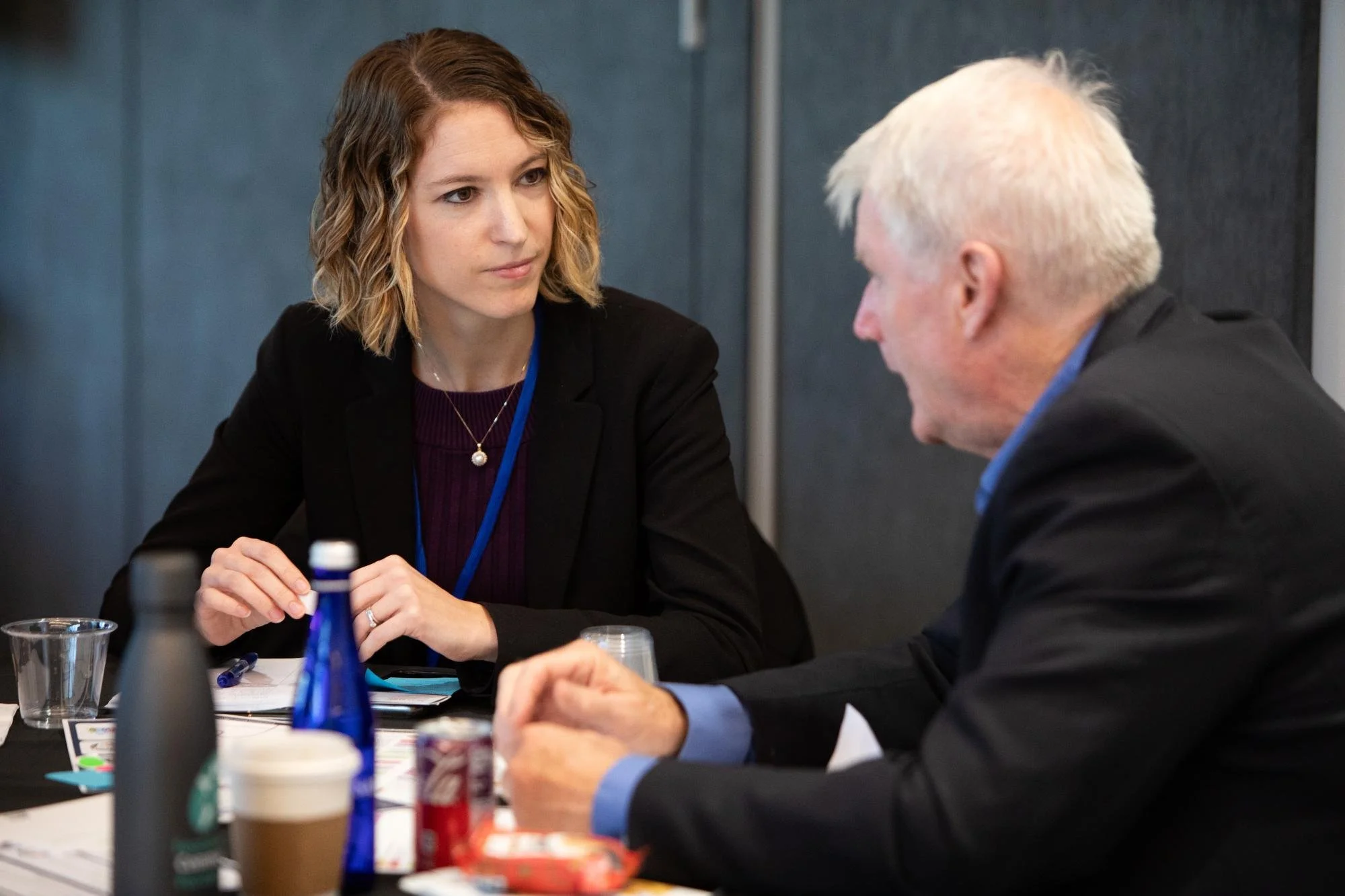 A woman with wavy, shoulder-length hair and a woman with short, white hair are sitting at a table, having a conversation. The table has bottles, cups, and papers on it, indicating a meeting or discussion.
