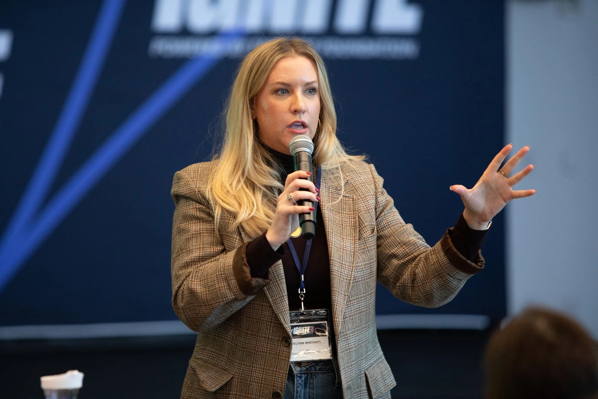 A woman with blonde hair wearing a plaid blazer speaks into a microphone during a conference, with a large screen and audience members in the background.