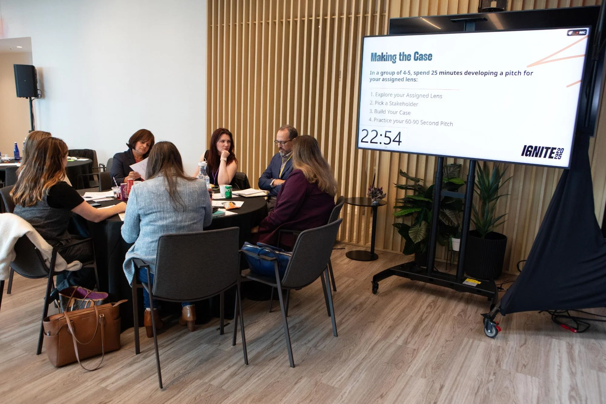 People sitting in a meeting room with a large screen displaying a presentation titled 'Making the Case' and a timer showing 22:54.