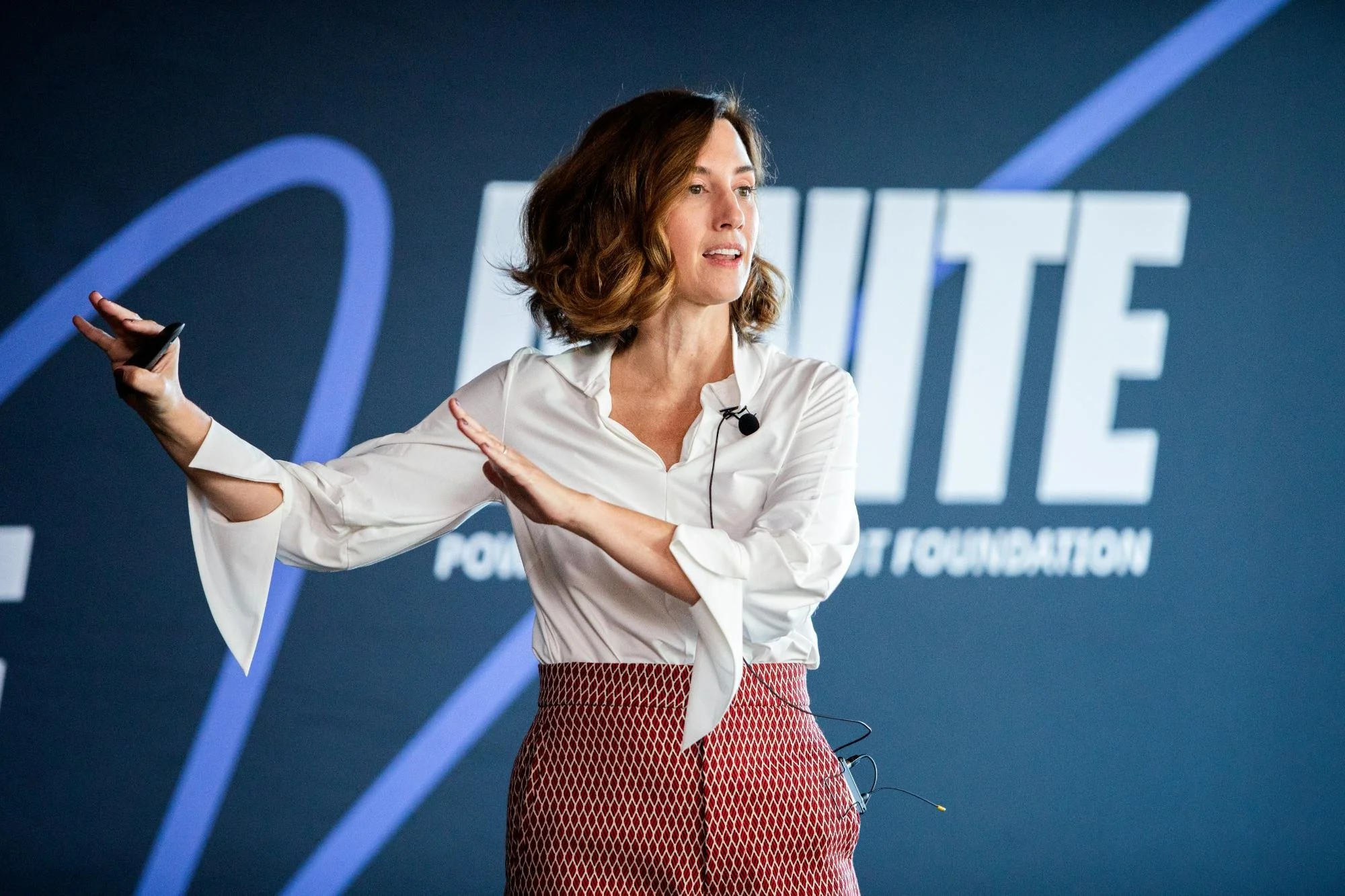 A woman with shoulder-length brown hair, wearing a white blouse and patterned red pants, is speaking or presenting at an event with a blue background and partially visible white text.