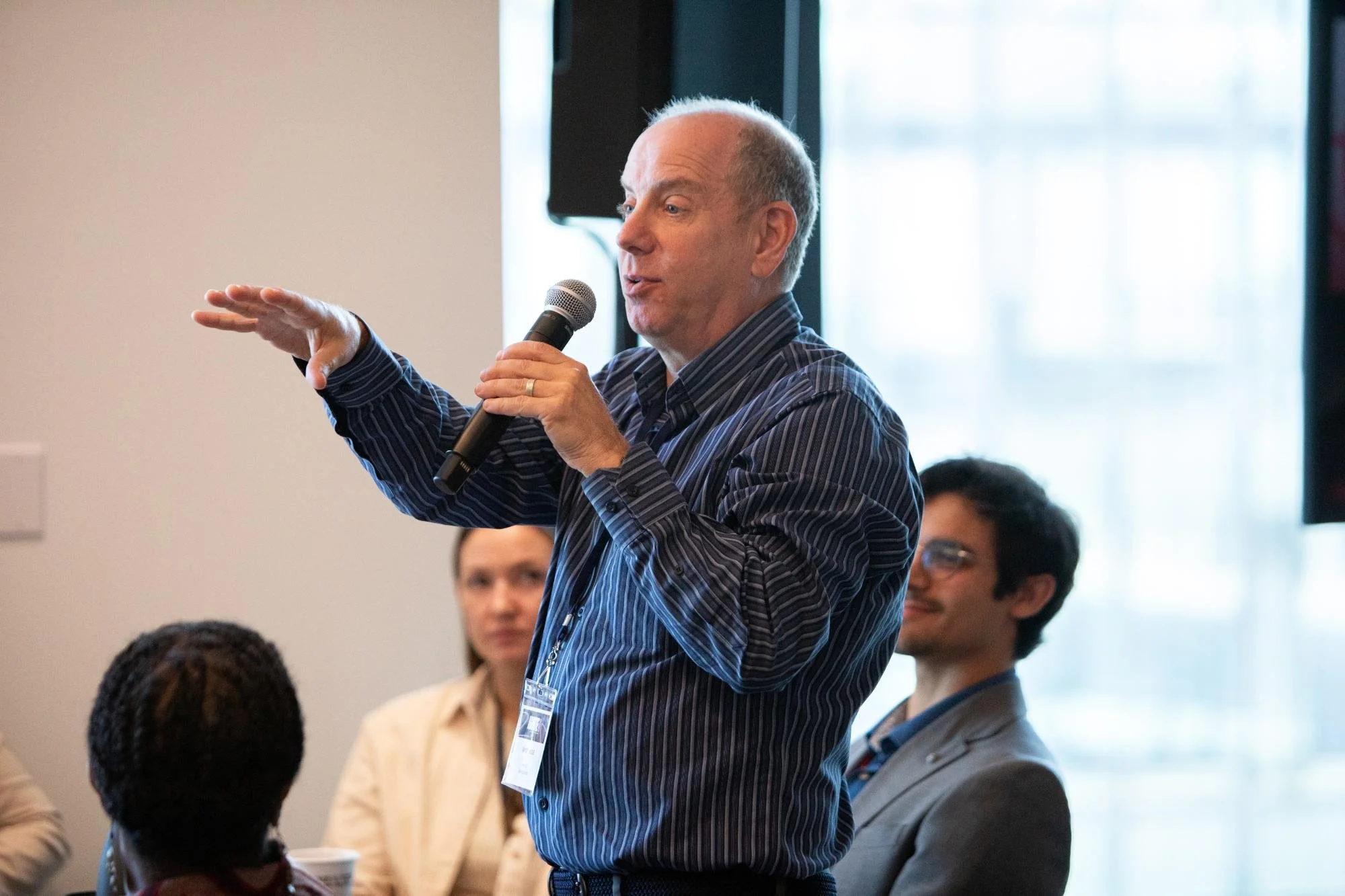 A man with a bald head and striped shirt speaking into a microphone during a panel or conference, with audience members seated around him and a large window in the background.