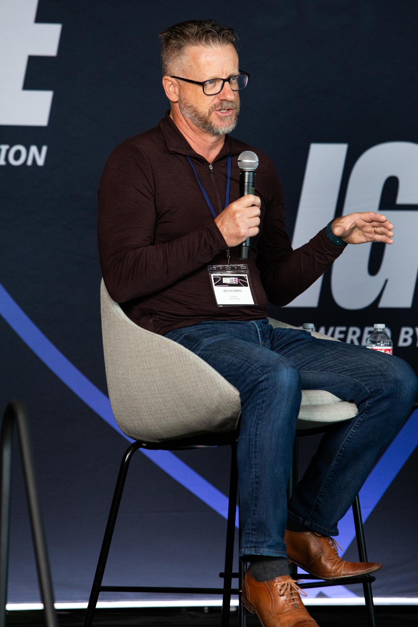 A man with glasses and a beard, wearing a black long-sleeve shirt, blue jeans, and brown boots, is sitting on a beige chair on stage, speaking into a microphone, during a panel or conference event.