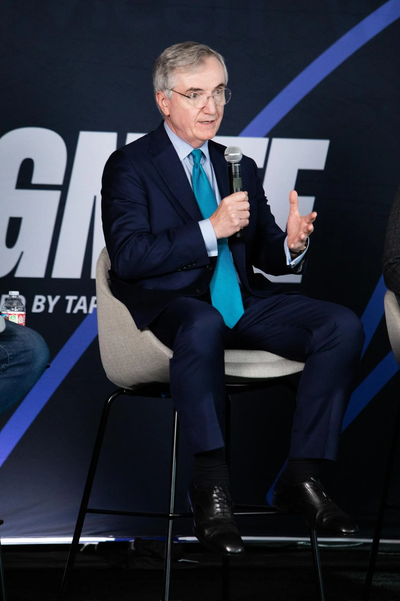 A man in a dark suit, light blue dress shirt, and teal tie sitting on a chair holding a microphone, speaking at an event with a black background and large white and purple text.