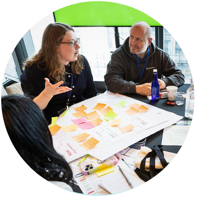A white woman gestures and speaks sitting at a table next to a bald man in a hoodie. In front of them is a large paper with sticky notes
