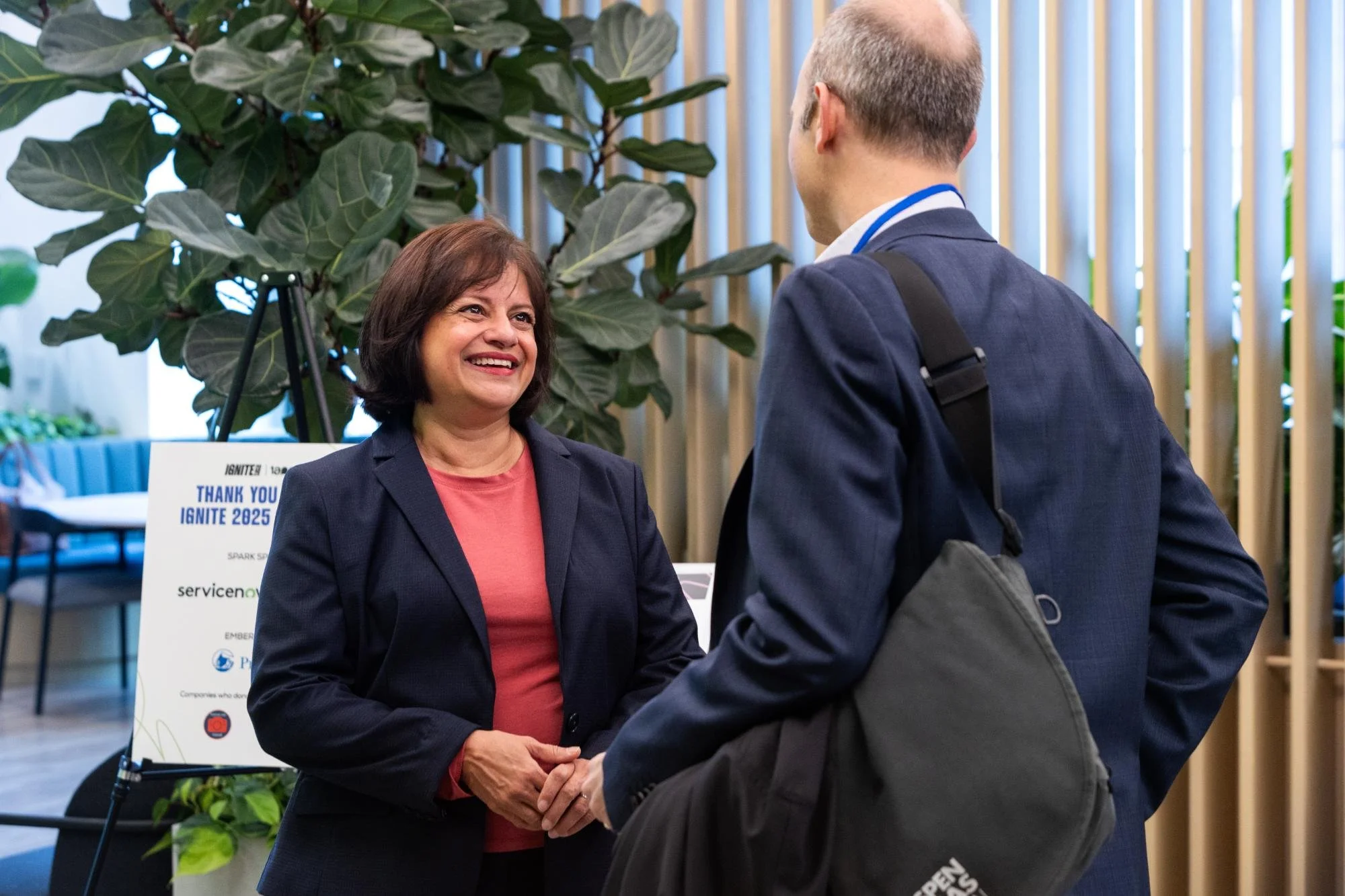 A woman with dark brown hair wearing a navy blazer and pink shirt smiling while shaking hands with a man in a suit and carrying a backpack, in a university or conference setting with large green plant and informational sign in the background.