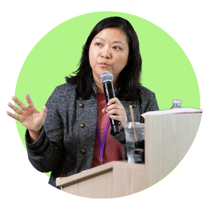 An Asian woman in a gray jacket speaks into a microphone and gestures while standing at a podium