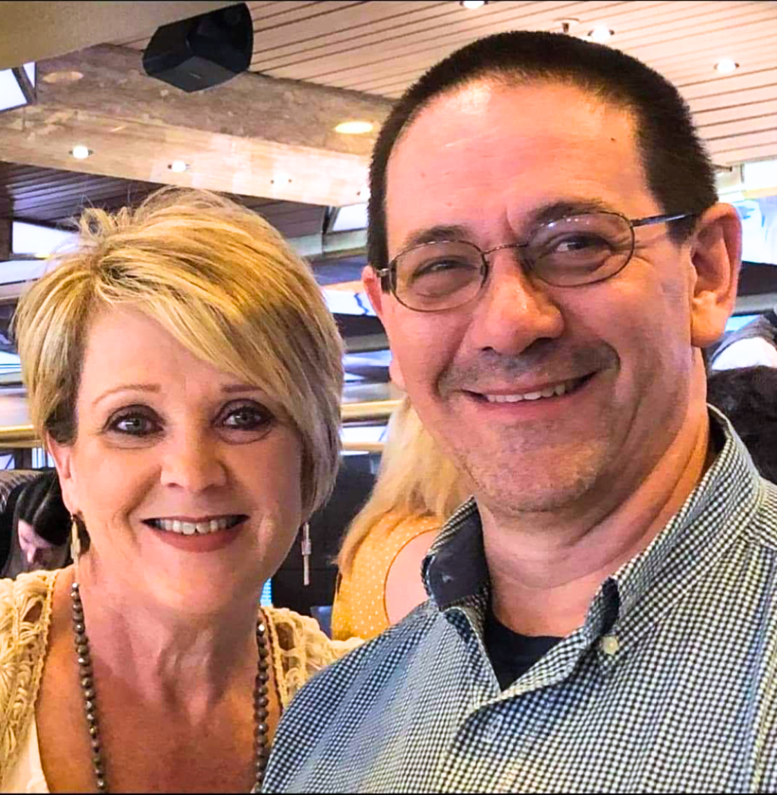 A smiling woman with short blonde hair wearing earrings and a necklace, and a smiling man with glasses and short dark hair, both taking a selfie at an indoor venue. These are the owners of AA Wholesale (Amory Auction), Doug and Angie Taylor.