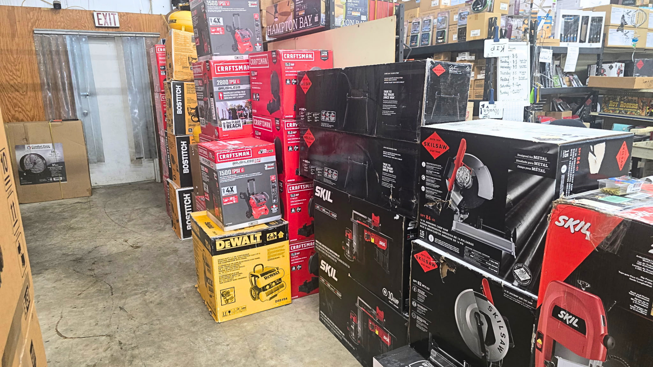 A store aisle with power tools and equipment in boxes, including chainsaws, drills, and blowers, stacked on shelves and on the floor. - AA Wholesale | Amory | MS | Wholesale | Deals | Amory Auction