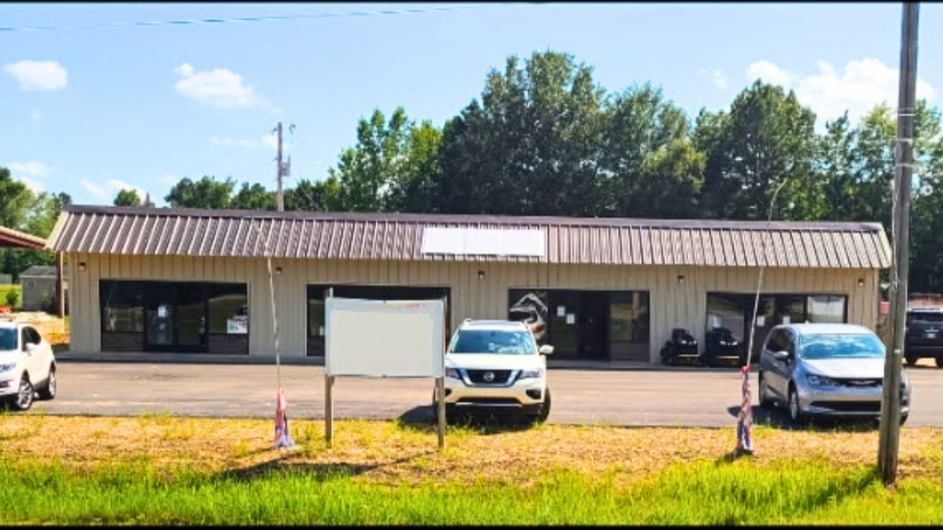 Single-story commercial building with a metal roof and large glass windows, parked cars in front, and trees in the background. - AA Wholesale | Amory | MS | Wholesale | Deals | Amory Auction