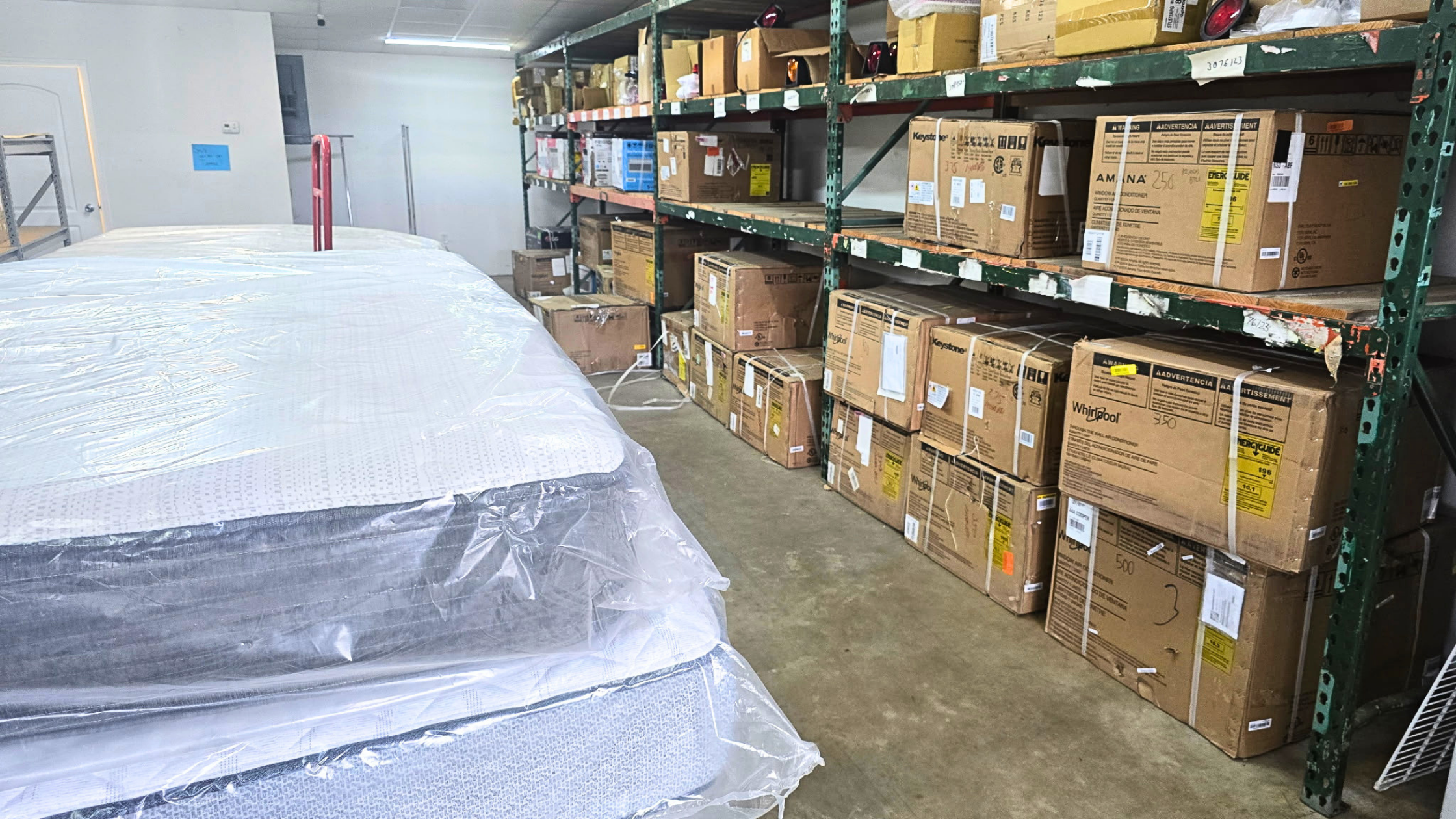 Warehouse storage area with stacked boxes on metal shelves and a large wrapped mattress or bedding item in the foreground. - AA Wholesale | Amory | MS | Wholesale | Deals | Amory Auction