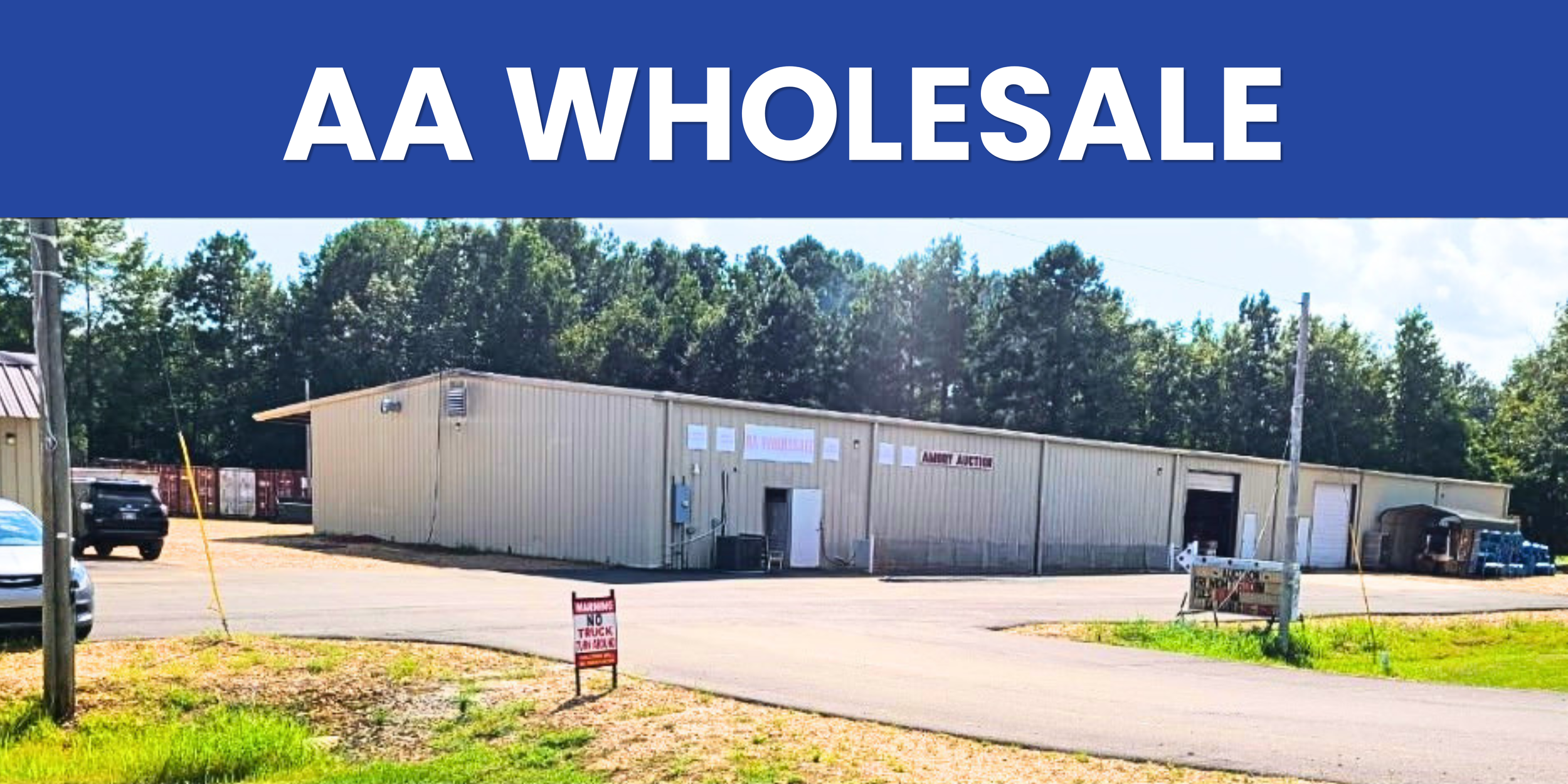 Large beige building with signs indicating it is an auction house, surrounded by trees and parked vehicles. A 'No Trucks' sign is visible in the foreground. - AA Wholesale | Amory | MS | Wholesale | Deals | Amory Auction