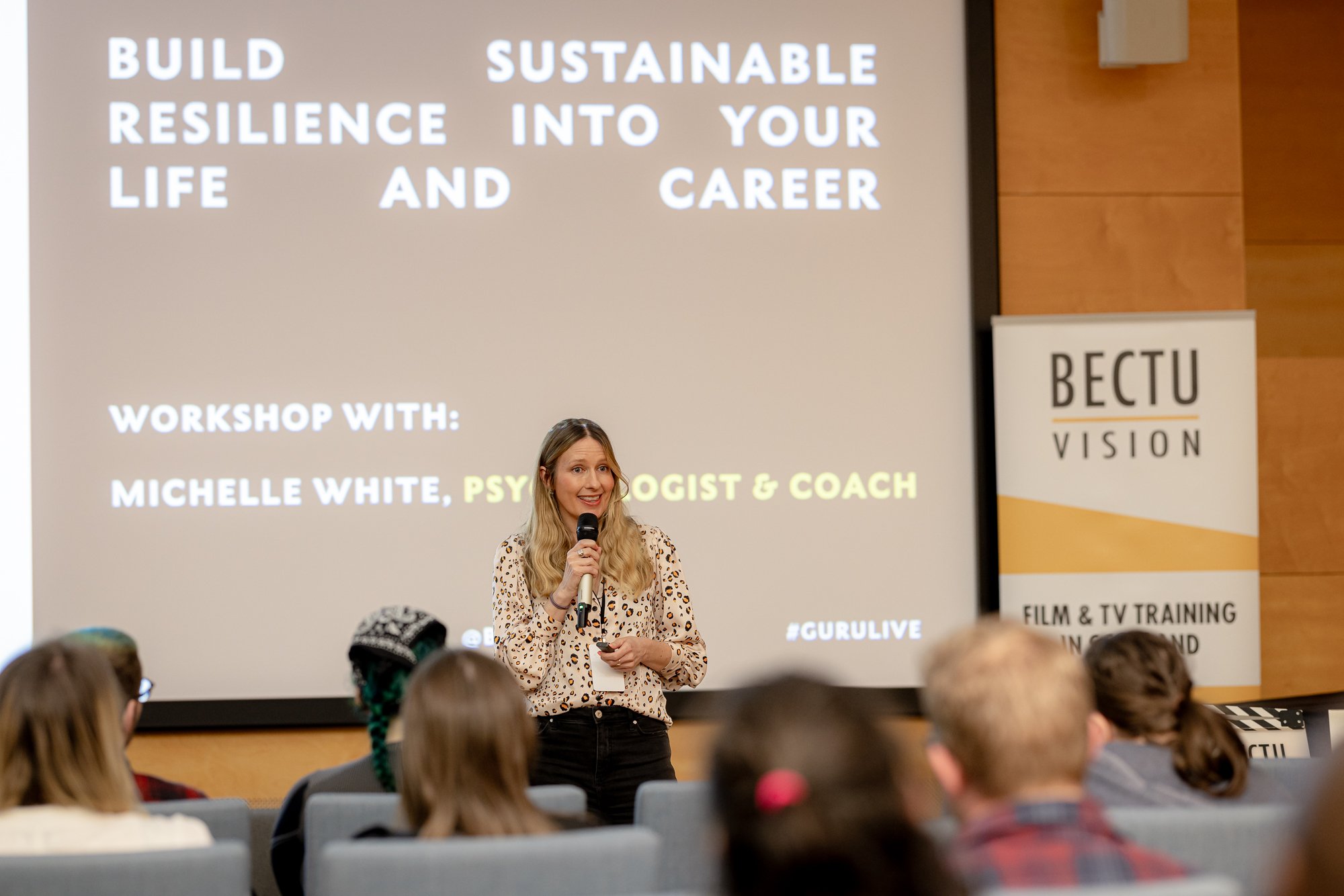 Woman with long blond hair speaking into a microphone in front of an audience at a workshop, with a large presentation screen that reads "Build Resilience into Your Life and Career."